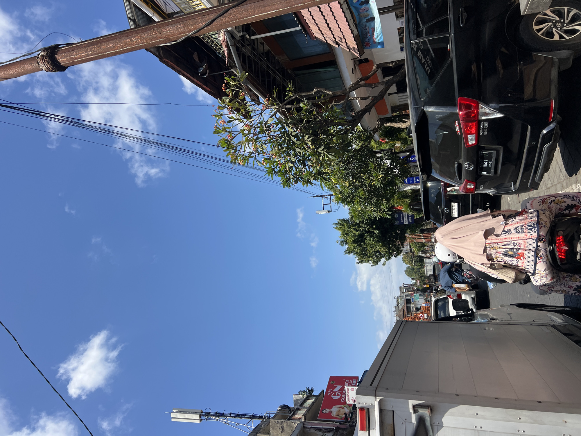 The image captures a street scene in Denpasar, Indonesia, under a clear blue sky with scattered white clouds. The perspective is from a vehicle in traffic, looking forward and slightly upwards. In the foreground, a white box truck is partially visible on the left. A person on a scooter, wearing a light-colored headscarf and a patterned dress, is in front of the truck, facing away from the camera. Behind the scooter, a black SUV is prominent, with its rear visible. Further down the street, other vehicles, including cars and possibly a motorcycle, are present, indicating traffic congestion.
On the right side of the image, buildings line the street. One building has a rustic, weathered utility pole in front of it, adorned with numerous wires. This building features a multi-level structure with awnings and balconies. A tree with green leaves and some pink flowers is situated in front of this building. Another building to the left appears to be older, with a visible cell tower on its rooftop. A red billboard with text and an image of a person is mounted on its side. In the background, more buildings and trees are visible, suggesting a commercial or residential urban area. The overall scene conveys a typical day in a busy Indonesian city with significant vehicular traffic.