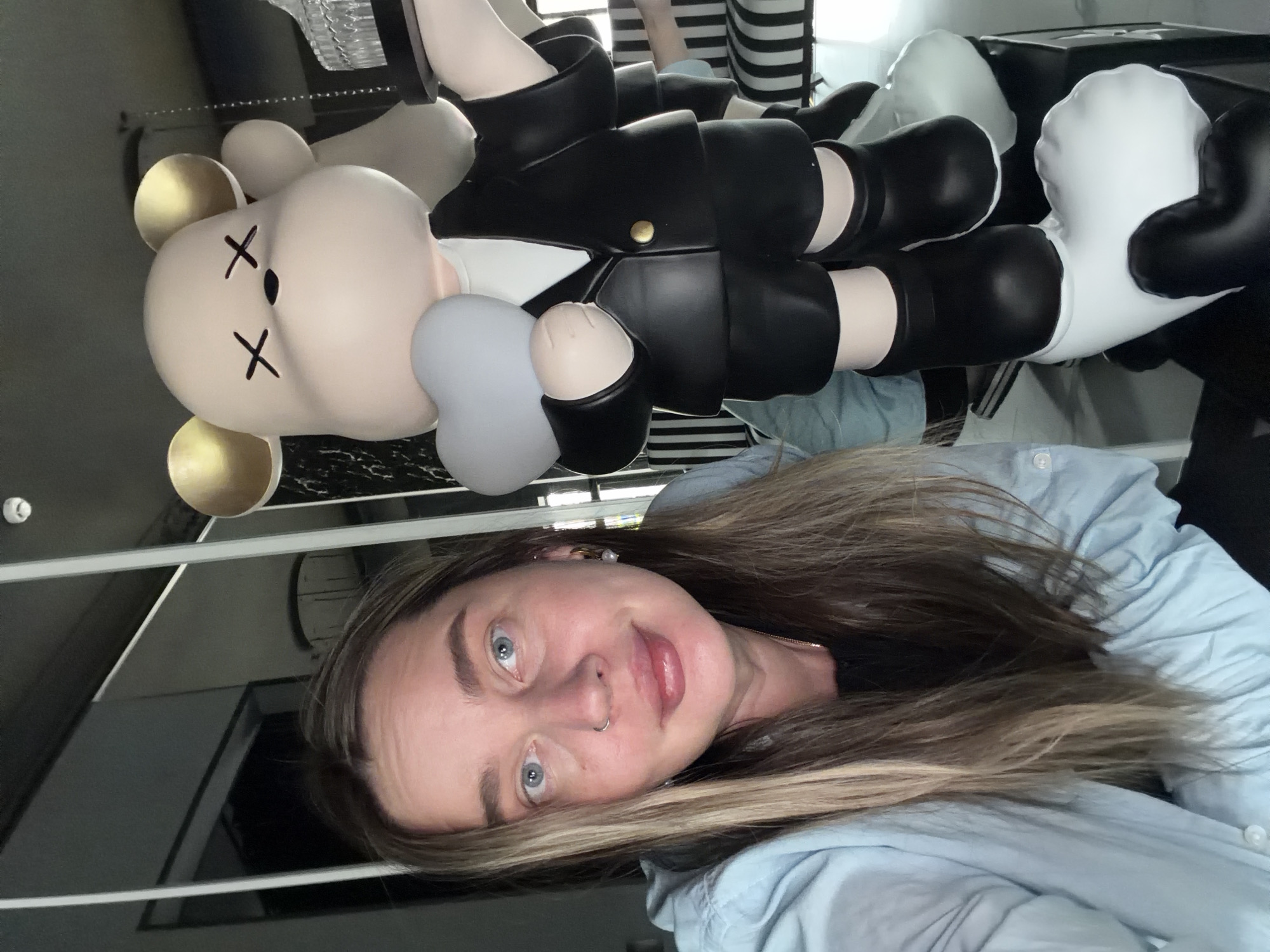 A woman with long, light brown and blonde hair is taking a selfie indoors. She is wearing a light blue button-down shirt. Behind her is a stylized bear figure with X's for eyes, wearing a black suit. The figure is hanging, possibly from a ceiling. Striped patterns are visible behind the bear, suggesting furniture or a wall design. The background has hints of what could be a modern interior design with dark accents. There is no definitive evidence indicating this photo was taken in Moscow, Russia.