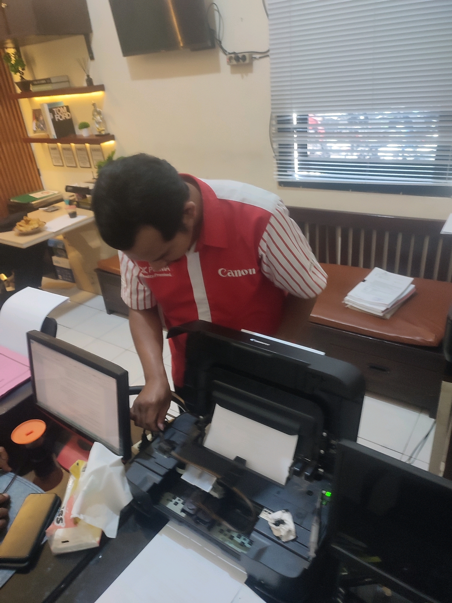 In Ringinanom, Indonesia, a man wearing a red and white striped polo shirt with "Canon" and other branding visible on it is seen tending to an open printer. The printer is black and appears to be a multi-function device. The man is leaning over the printer, with his hands engaged in what seems to be maintenance or repair. In the background, a desk with a computer monitor, a phone, and a box of tissues are present. Further in the background, a shelf displays books, including one with "TOM FORD" visible, and decorative items. The environment suggests an office or home office setting with a window covered by blinds and a wooden bench.