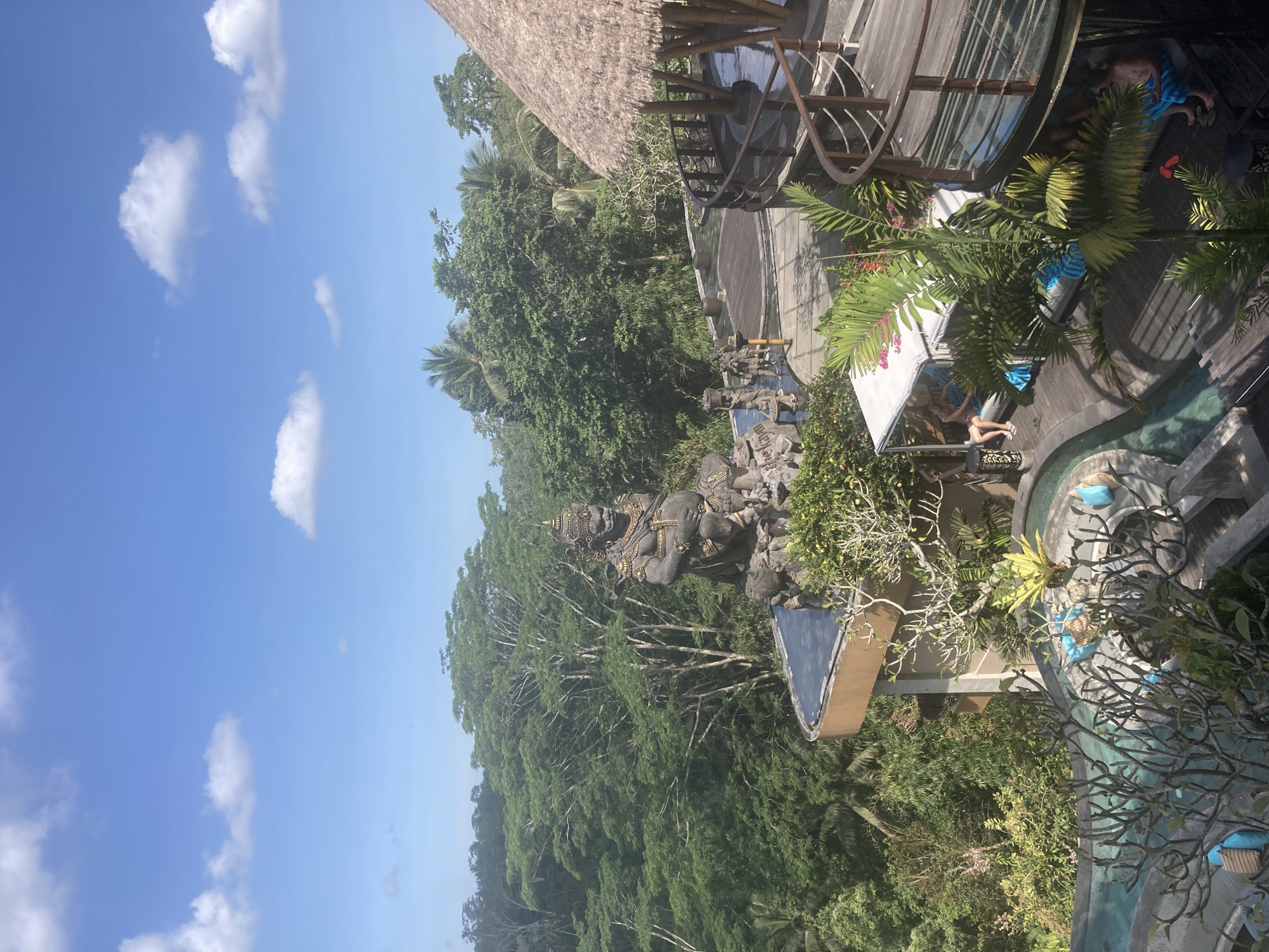 The image shows a view of a tropical landscape with a building featuring a thatched roof and wooden structure. There is a statue amidst lush greenery. Some people are visible on the wooden deck. The location is Unknown, Unknown.