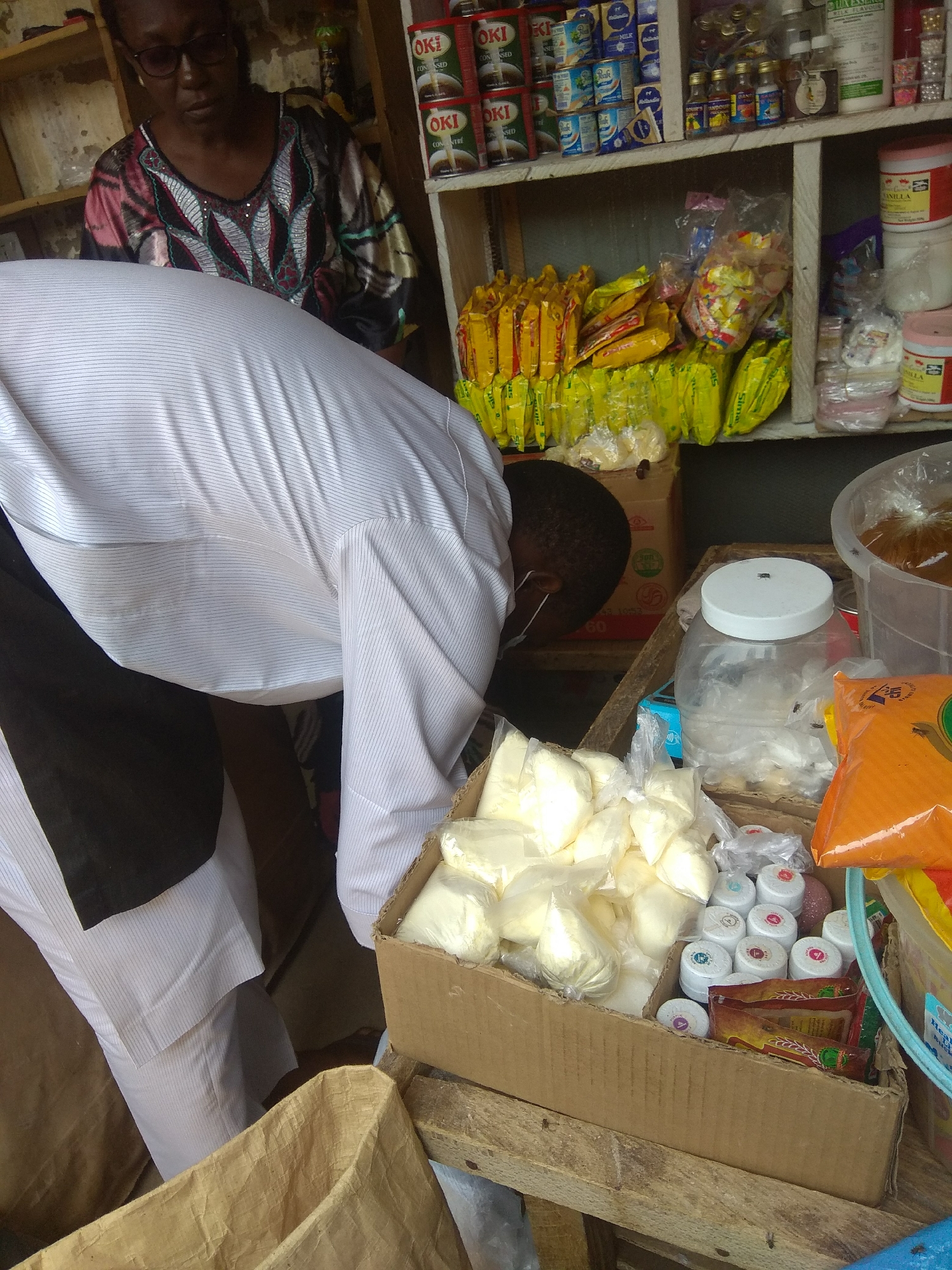 This is a medium shot of a person, likely a vendor, bending over a display of goods in what appears to be a market stall. The person is wearing a white pinstripe shirt and dark pants. Behind them, a woman with glasses, wearing a colorful patterned top, is partially visible.
The stall is filled with various items, including packaged food products like cans of "OKI" condensed milk, yellow packets of snacks, and small bottles and jars of other consumables. A large cardboard box in the foreground contains numerous small, individually wrapped white packets, possibly spices or powders, alongside small containers with colorful lids. A large plastic jar with a white lid, containing some white material, is also prominent, with several flies visible on and around it. To the right, a large clear container holds a brown granular substance, and a large orange packet is also visible. The shelves are wooden, and the overall impression is of a bustling, possibly outdoor, market. The lighting suggests daytime. The visible text indicates brands like "OKI," "MILK," and "DANILLA," and hints at the location with "Jalingo, Nigeria" in the metadata. The scene conveys a sense of commerce and daily life.