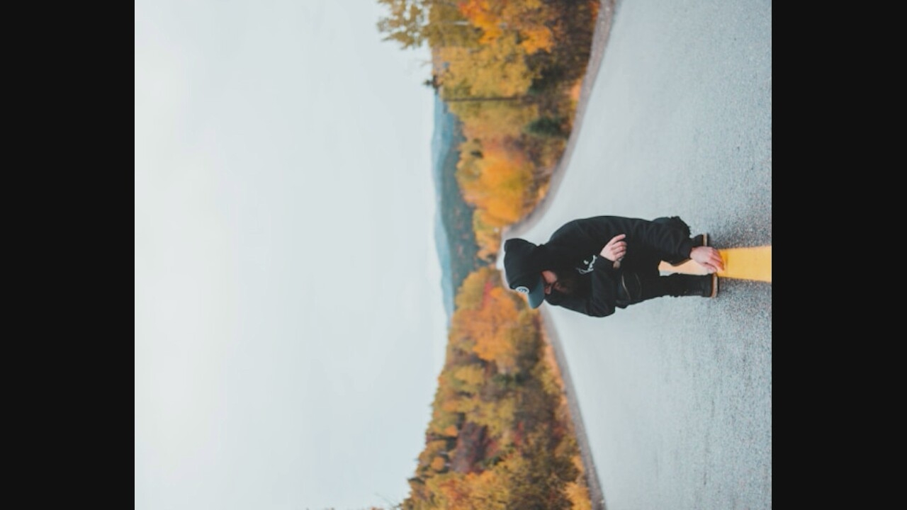 An individual wearing a black hooded sweatshirt and a black baseball cap is crouching on a wet asphalt road in the middle of autumn. The individual has a beard and is looking to their left. A yellow line is painted down the center of the road, and the individual is positioned with their hands resting on it. The background shows a landscape of trees with colorful fall foliage, ranging from yellow to orange and red, with a hazy sky above. The scene appears to be in a rural or mountainous area.