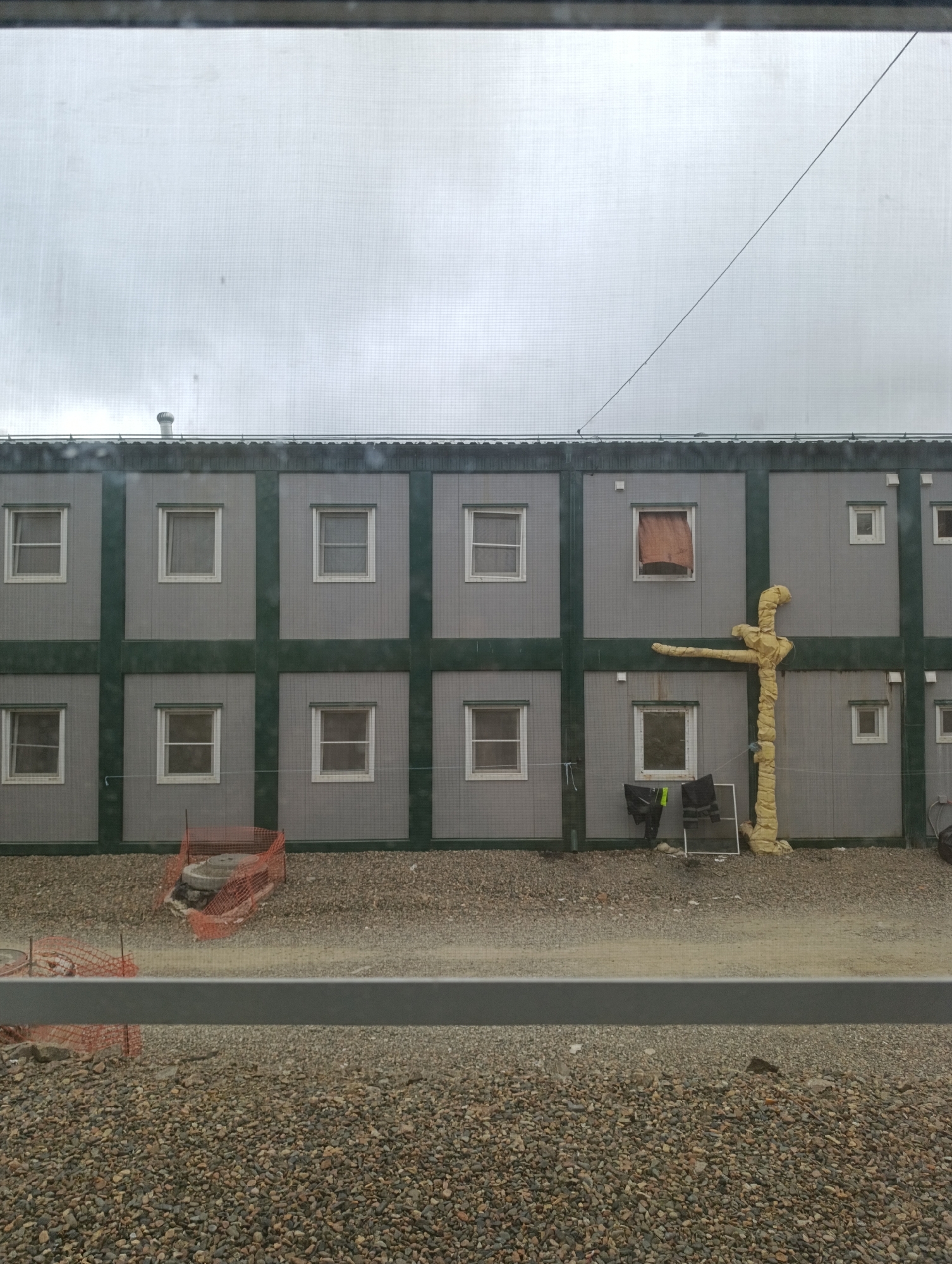 The image depicts a row of prefabricated modular buildings, likely temporary housing or office units, viewed from behind a window with a visible screen. The buildings are gray with dark green trim, featuring a grid pattern of identical square windows. A gravel-covered ground is visible in front of the buildings, with some construction barriers and materials.

To the right of the building's center, an unusual yellow, pipe-like structure extends horizontally from the building, with a vertical section wrapped in insulation material. Below this, a pair of dark pants and a yellow vest are hanging on a line, along with a white frame structure.

The sky is overcast and appears to be a light gray. The image quality is slightly obscured by the window screen. The scene suggests a construction or work site in an outdoor environment, possibly in Kurleya, Russia, based on the provided location context. There are no visible people in the image.