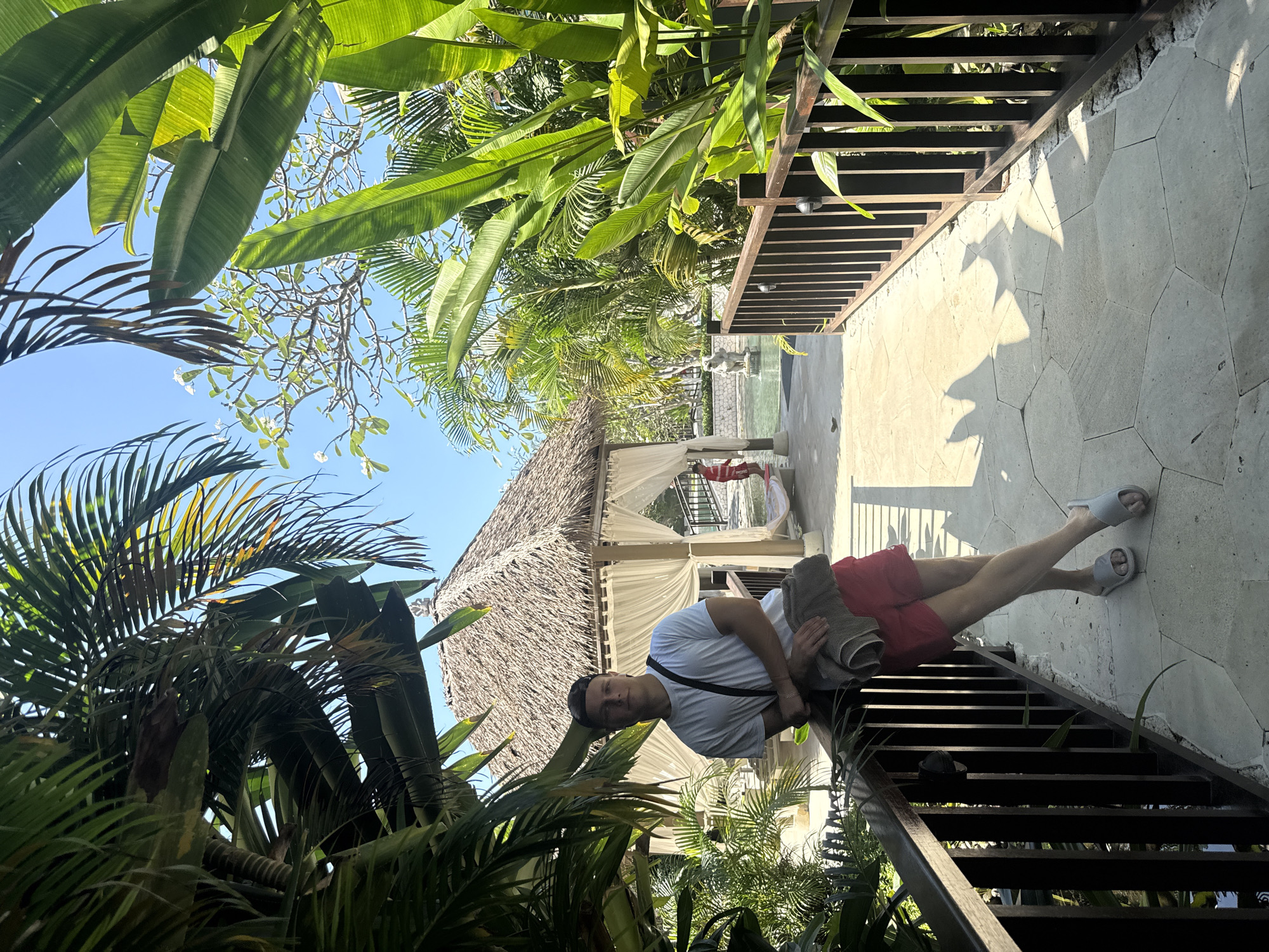 A medium-shot image is taken at an oblique angle, showcasing a man standing on steps that appear to lead away from a cabana and towards an open area. The likely location is Kuta Selatan, Indonesia, based on the tropical flora and architecture.
The man is dressed in a white t-shirt and red shorts, wearing sandals. He has a black strap across his chest and is holding a rolled-up towel. The stairs he stands on are dark with horizontal treads, lined with a metal railing.
The backdrop includes a structure with a thatched roof and white curtains, possibly a cabana. A red hammock is visible inside. The foreground and background feature lush tropical vegetation, including palm trees.
Above, there is a clear blue sky, with some foliage partially obscuring the view. The ground is paved with stone. Light suggests it is daytime.