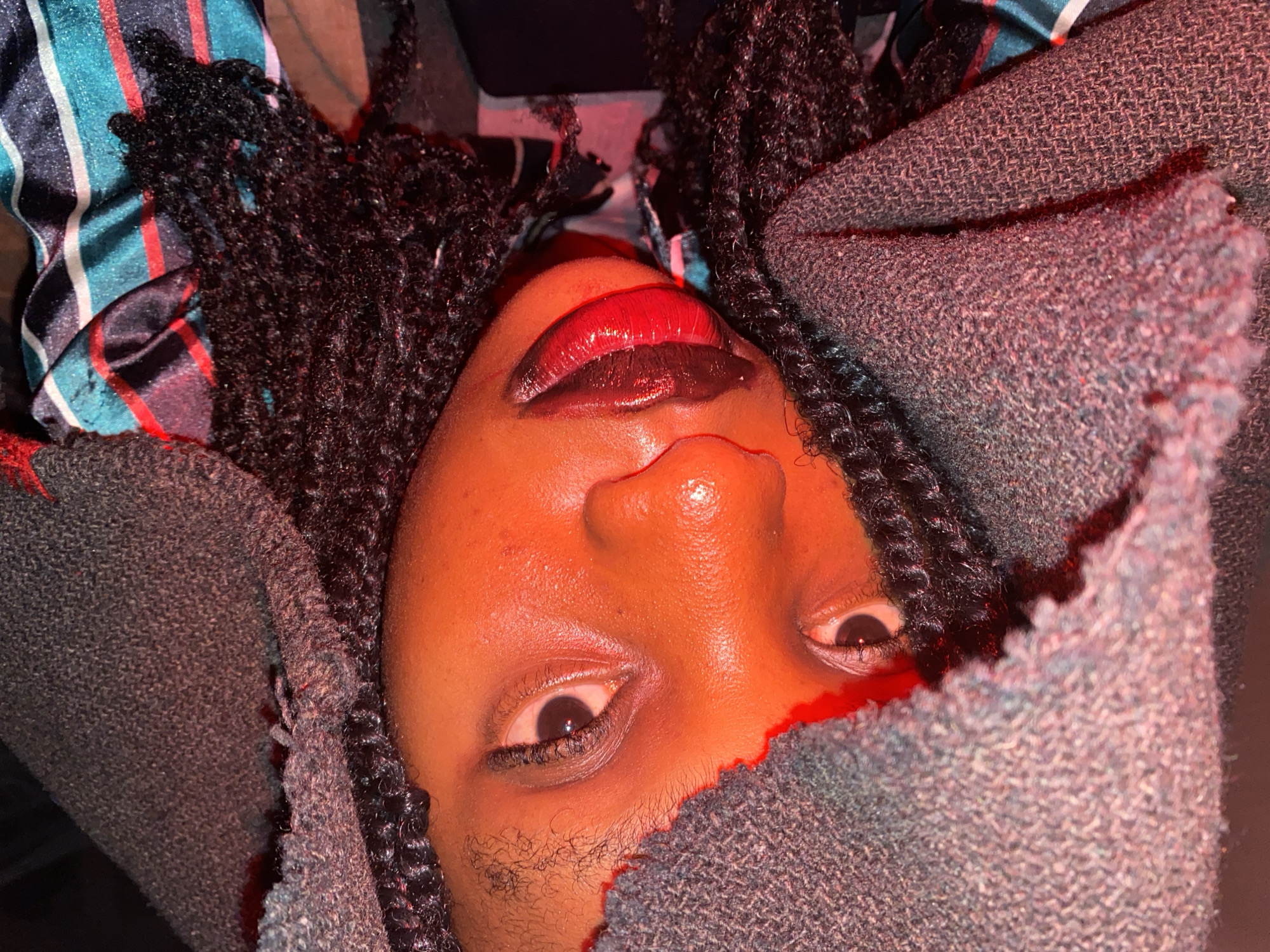 A close-up, upward-facing shot features a person's face surrounded by elements of fabric and hair. The individual has dark skin, with visible facial hair above the lip. Their lips are accentuated with a two-tone makeup, with a red hue on the upper lip and a darker shade on the lower. The individual's dark eyes are visible, gazing upwards. Their hair is styled in braids. A grey fabric texture is draped on either side of the face, partially obscuring the forehead and chin. A different fabric, with a striped pattern of blue, red, and white, is visible at the top of the frame. The lighting is uneven, with a reddish cast on the face. The city is Unknown, Unknown.