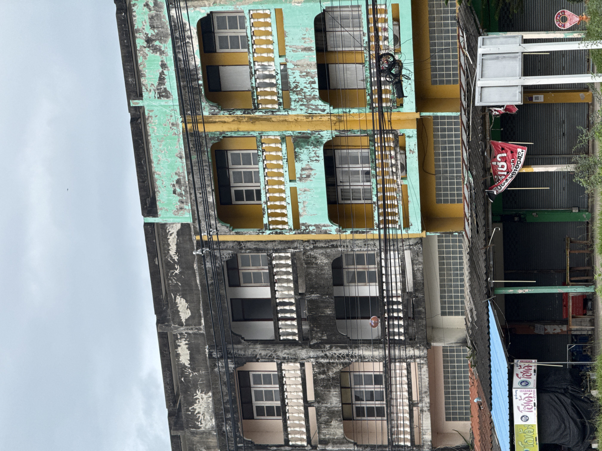 The image depicts the weathered facades of two adjacent multi-story buildings in Ban Nong Bua, Thailand. The building on the left is predominantly dark gray with signs of decay and water stains, featuring light pink balconies with white balustrades and white-paned windows. The building on the right is painted in a vibrant, yet peeling, turquoise and yellow color scheme, with similar balconies and windows. Overhead, a dense network of black electrical wires crisscrosses the scene. Below the upper floors, a covered area with various shopfronts is visible. These shops have corrugated metal roofs, some with blue awnings, and metal roll-down shutters. Signs with Thai script are displayed, and some shops appear to be closed. A red banner with Thai writing hangs from one of the shops. The ground in front of the shops is covered in low-lying vegetation and grasses. The sky is overcast with a dull, gray hue.