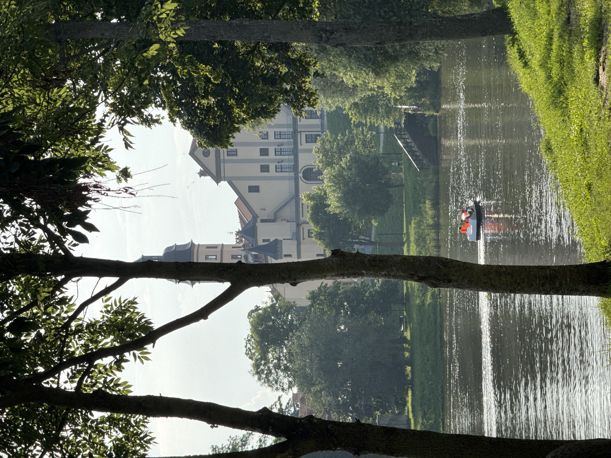 This is a full-frame, vertical, outdoor, eye-level, daylight shot of a scenic view overlooking a body of water with a castle-like building in the background. In the foreground, two large tree trunks frame the scene, with lush green grass and foliage to the right. The water in the middle ground is calm with small ripples and reflections of light. A white pedal boat with four people wearing orange life vests is moving across the water. In the background, a grand, light-colored building with a central tower and multiple windows is visible on a slight incline. Trees and greenery surround the building and line the banks of the water. A pedestrian bridge with dark railings is situated on the right side of the water, leading towards the building. Several people are walking on the path near the bridge. This scene is of the Nesvizh Castle complex in Nesvizh, Belarus.