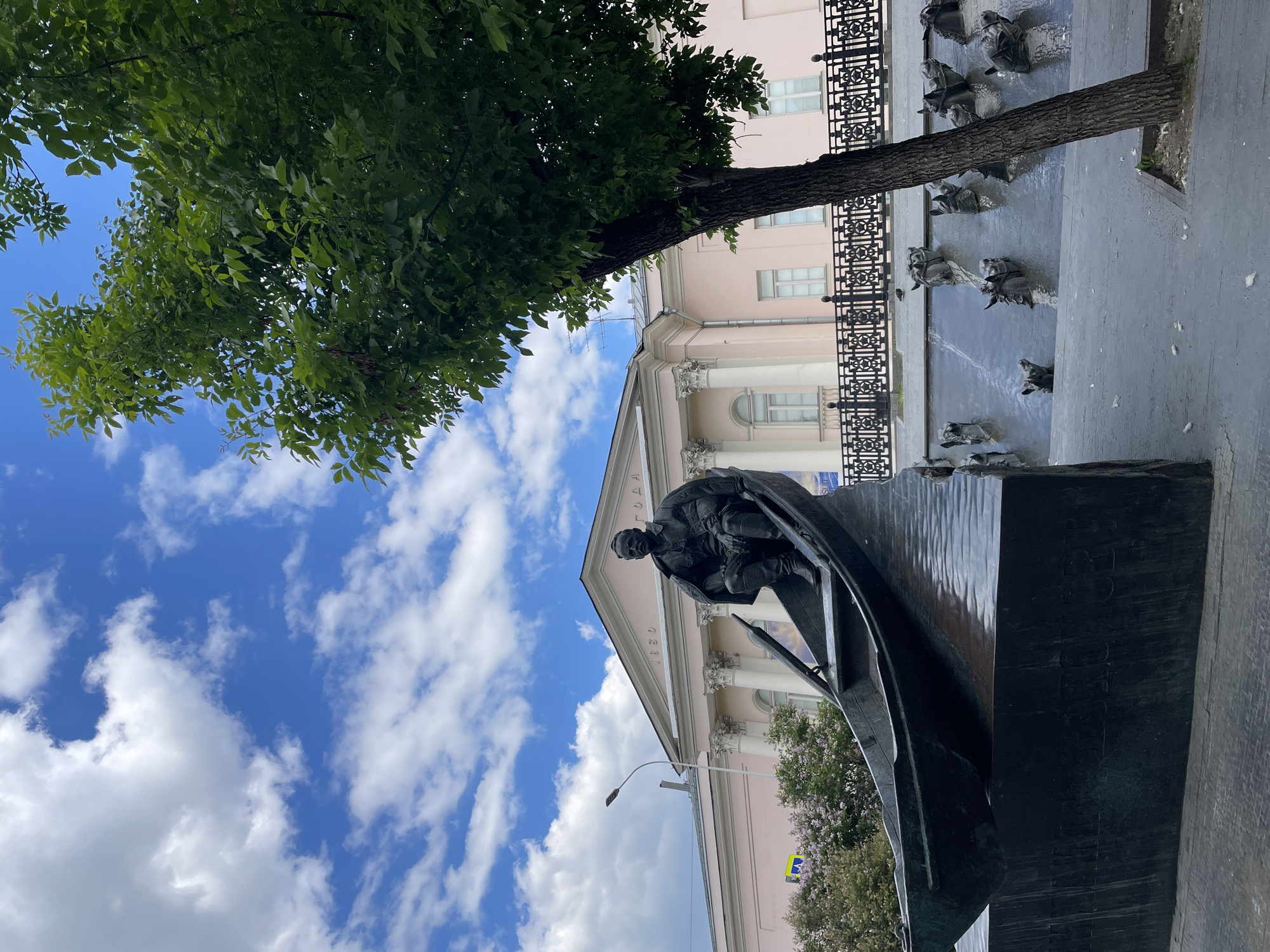 A striking vertical shot captures a public square in Moscow, featuring a prominent dark statue against a backdrop of classical architecture and a bright sky.
In the foreground, a large, dark-colored bronze statue depicts a seated male figure in historical attire within a stylized boat-like structure. The figure appears to be a historical personality or writer, positioned dynamically. This main statue stands on a dark, textured pedestal.
Behind the statue, to the right, a black metal fence with intricate decorative elements separates the foreground from a larger sculptural installation. This installation features multiple smaller, dark figures, possibly busts or heads, emerging from a dark, wavy surface, reminiscent of a fountain or a memorial.
The background is dominated by a grand, light pink or beige building exhibiting classical architectural features, including prominent white columns and a balustrade. Above the columns, text is visible, including the Cyrillic word "ГОГОЛЯ" (Gogolya) and the year "1830", indicating the monument is dedicated to Nikolai Gogol and the building might be associated with him or this period. A street light pole is also visible.
A large, leafy green tree occupies the upper left portion of the image, its dense foliage creating a natural frame. The sky is bright blue with scattered, fluffy white clouds, suggesting clear, sunny daytime weather. The image composition, with its high angle, emphasizes the monumentality of the statue and the architectural setting.