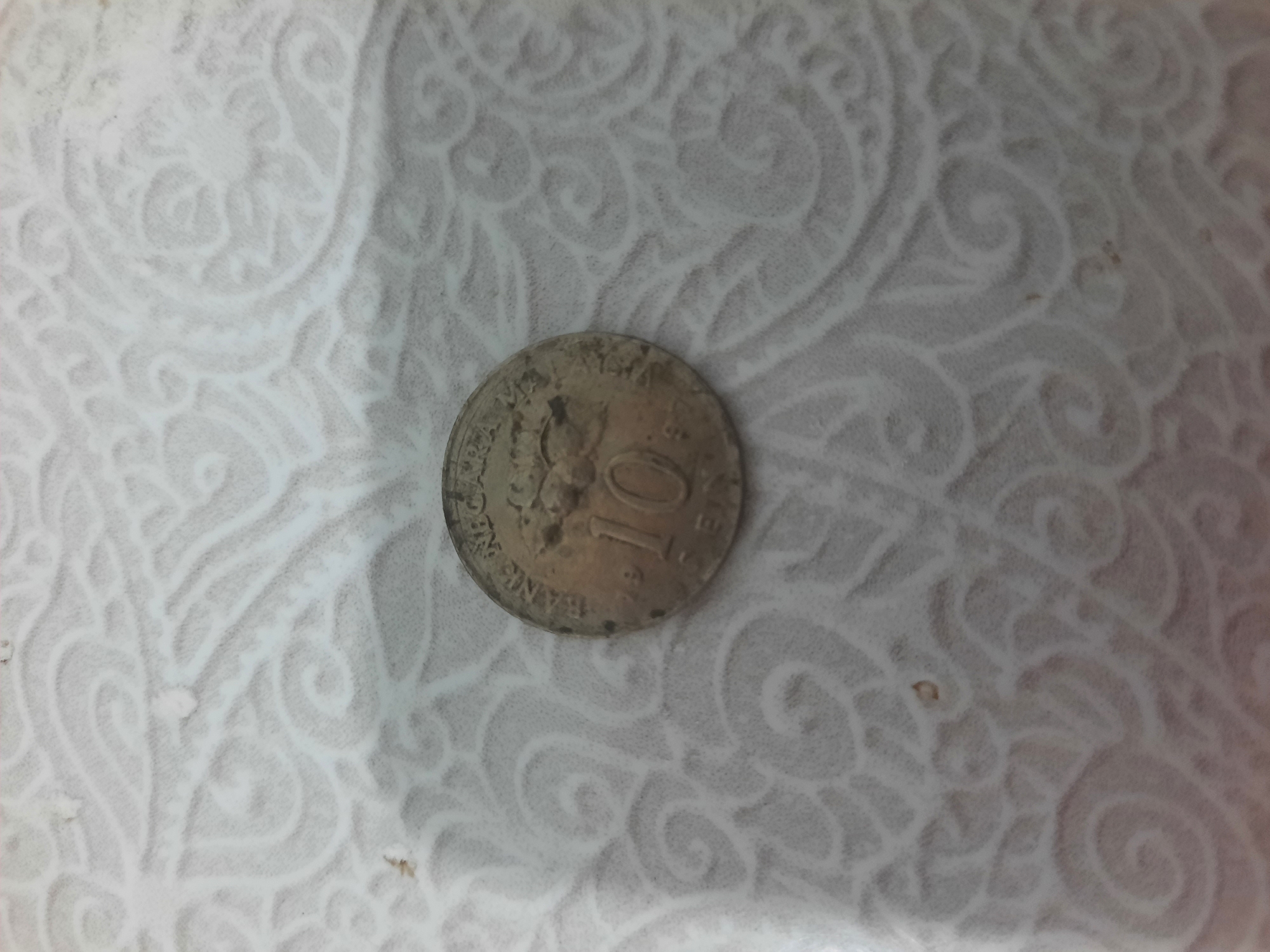 A close-up, overhead shot of a worn, tarnished coin rests on a white textured surface. The coin is silver in color and has visible wear and tear. The face of the coin displays the number "10" prominently, with the inscription "BANK NEGARA" above it. Below the "10", the word "SEN" is visible. The coin appears to be from Indonesia. The textured surface beneath the coin has a repeating, intricate pattern of swirling lines. There is no visible context to suggest the city of Lhokseumawe, Indonesia.