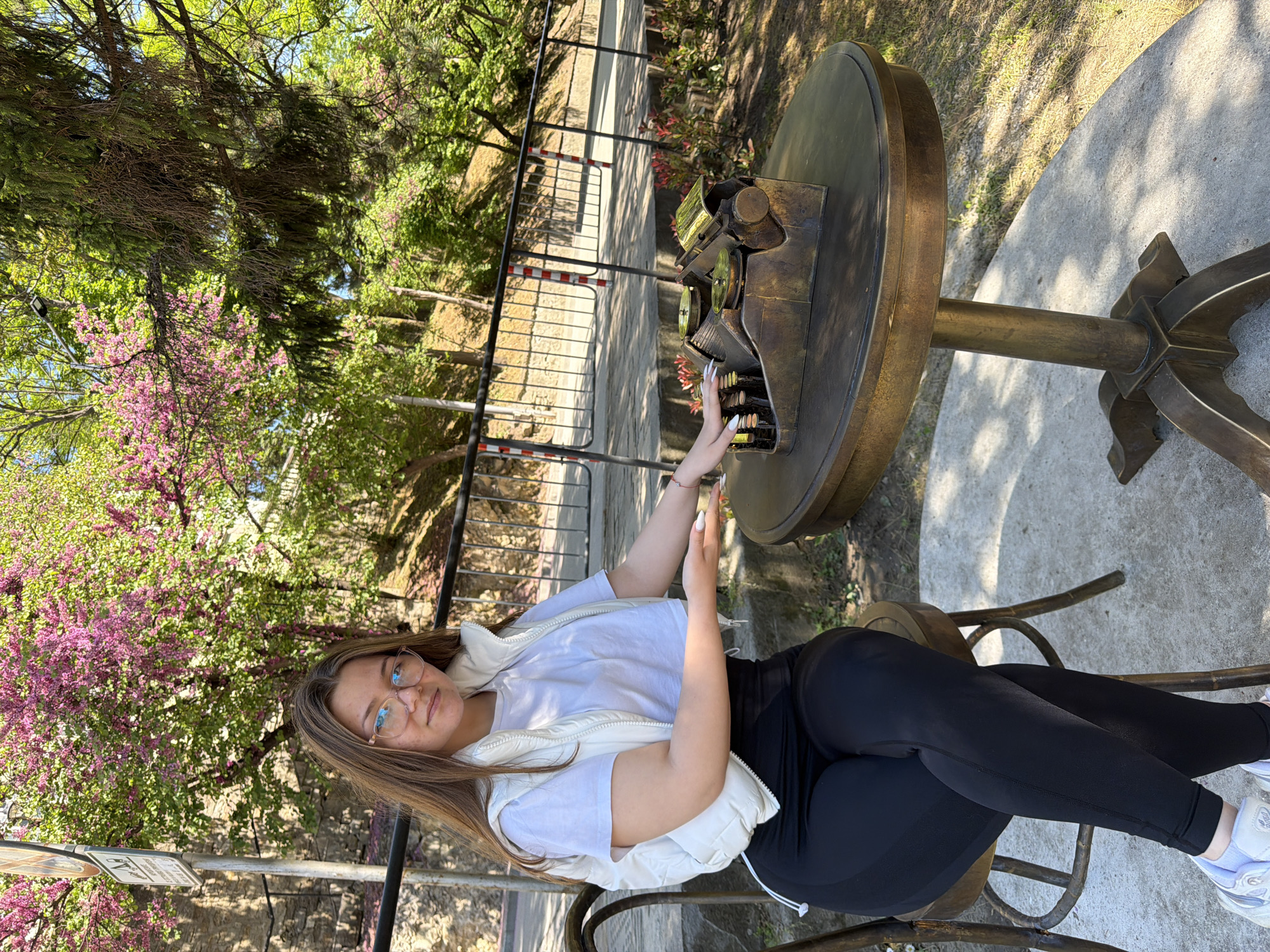 The image depicts a young woman seated outdoors in what appears to be a park or garden setting, likely in Veliko Tarnovo, Bulgaria. She is wearing eyeglasses, a white t-shirt, a light-colored vest, and black leggings, with white sneakers visible on her feet. She is posed next to a unique, possibly bronze or metal table that features a large, ornate representation of a typewriter on its surface. Her hands are near the keyboard of the typewriter sculpture. The surrounding environment includes trees with pink blossoms and green foliage, along with a black metal fence in the background. The ground appears to be paved or stone.