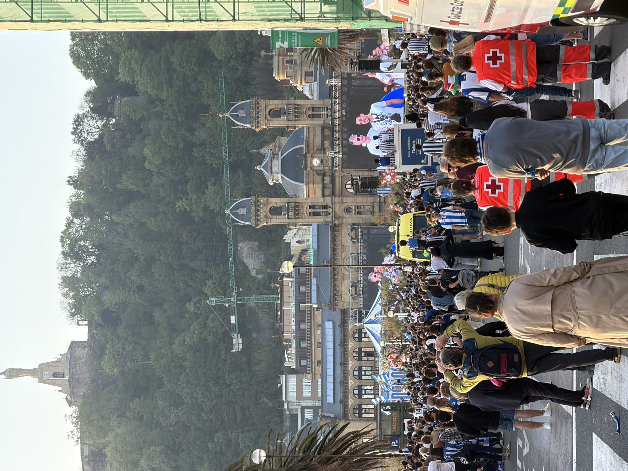 The image, when correctly oriented, captures a vibrant public gathering on a sunny day in San Sebastián, Spain. A dense crowd of people fills the foreground and mid-ground, with many individuals wearing blue and white striped shirts, indicative of support for the local football club, Real Sociedad. The atmosphere suggests a celebratory event or parade.
In the immediate foreground, several individuals are visible from behind, including some wearing backpacks marked with a red cross, likely medical or first aid personnel. Two ambulances, one white and one yellow, are present within the crowd, positioned to the right and further back respectively. Large digital screens display images of people, suggesting a public viewing or live broadcast as part of the event.
The background showcases the distinctive architecture of San Sebastián, featuring ornate buildings with dark domes and intricate facades. Behind these structures, a large, lush green mountain rises, with a prominent statue of Christ (Sacred Heart) visible at its peak, characteristic of Mount Urgull or Igueldo. A green construction crane is also visible against the hillside.
Visible text elements include "TREN TURISTICO Tickets" on a sign, "ELAN" with a phone number on a banner, and "teipark" next to a parking "P" symbol on a street sign. The side of the white ambulance partially displays "Ambulancia / Ambulance." The clear sky and bright daylight indicate the event is taking place during the day, likely in the late morning or afternoon. Palm trees are also scattered throughout the urban landscape.