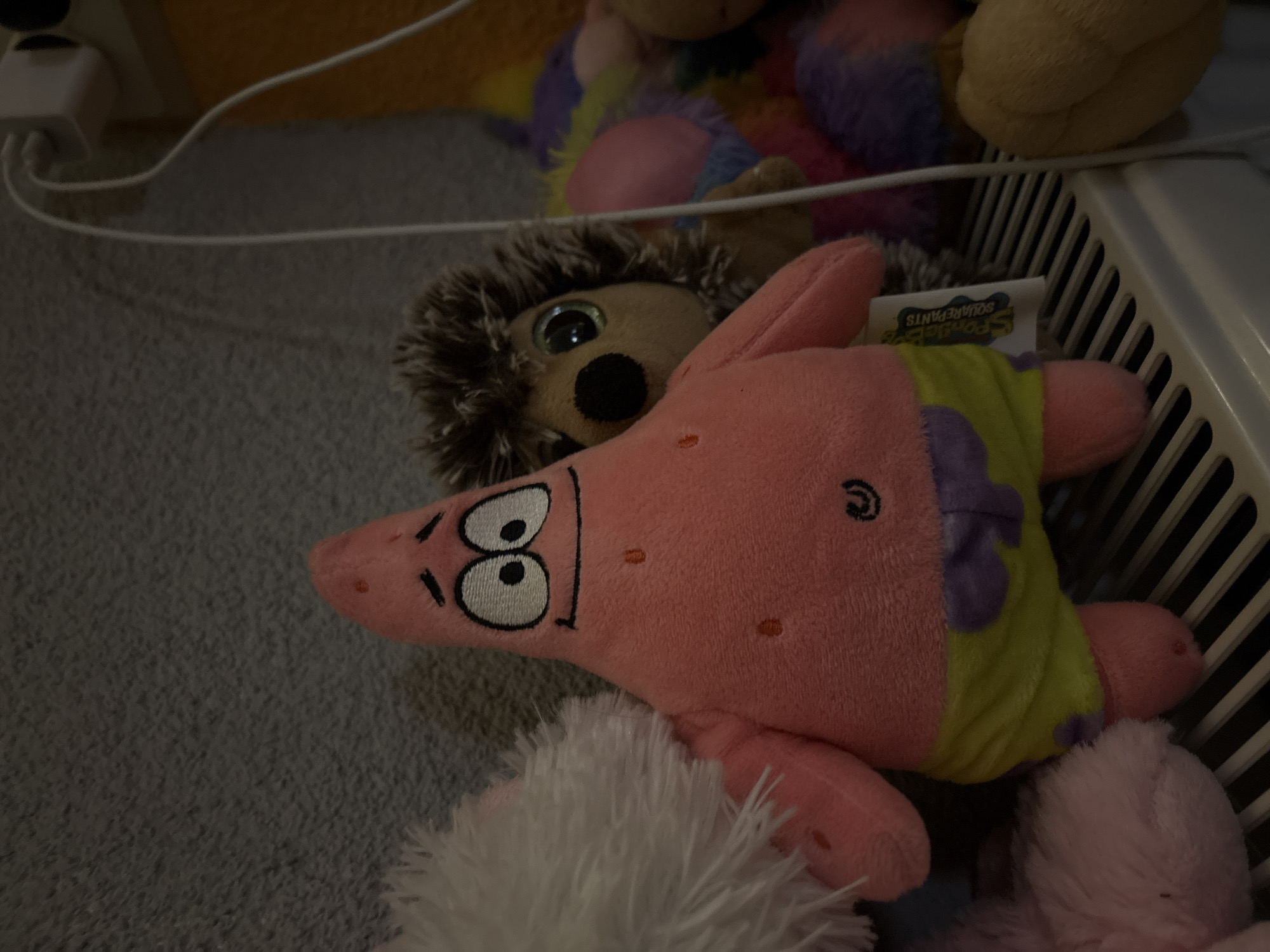 The image shows a close-up of several stuffed toys. A pink Patrick Star plush toy is in the foreground, with part of its face visible. Behind it is a brown hedgehog plush. A fluffy white stuffed animal is partially visible at the bottom of the image. A white power cable and wall plug are visible in the upper left. Other colorful plush toys, including a yellow one, are in the background. Some of the toys are partially within a white plastic container with vertical slats. There is no information that places it in Panagyurishte, Bulgaria.