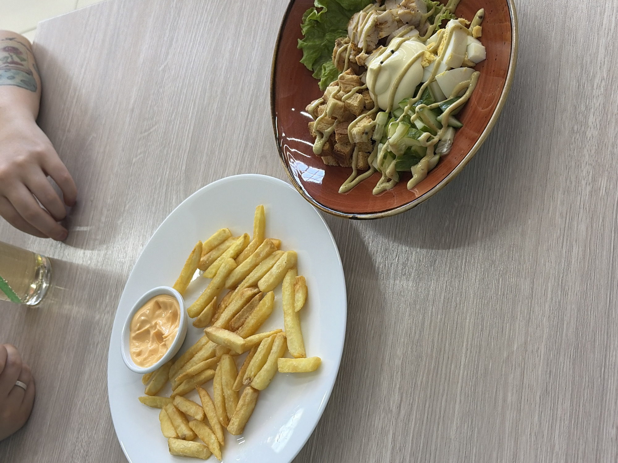 The image captures a dining scene, likely in a restaurant or café in Chaikovsky, Russia. Two plates of food are visible on a light wood-grained table. One plate, round and white, contains a serving of french fries and a small dipping bowl filled with a creamy, orange-colored sauce. The other plate, which is rounded with an orange rim, holds what appears to be a Caesar salad with grilled chicken, croutons, lettuce, sliced eggs, and a dressing. A person's arm with a visible tattoo is in the frame on the left side of the image. A glass of light-colored liquid is also present on the left side of the image. The composition suggests a top-down view of the table setting, with the focus on the two plates of food.
