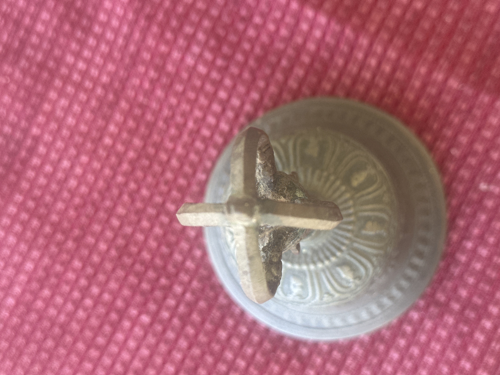 A close-up, overhead shot shows a small, antique-looking brass bell with a cruciform handle resting on a pink textured surface. The bell has decorative etching around its base. The pink surface appears to be a fabric with a raised, dimpled pattern. The image is taken from above the bell, with the bell occupying the bottom left quadrant of the frame, and the pink surface filling the rest of the image. There is no indication of the city of Thora, Australia in the image.