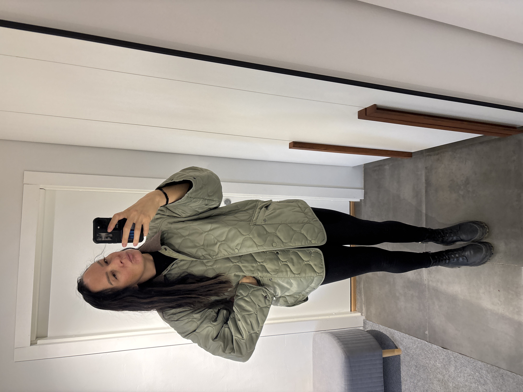 A woman stands in front of a mirror, taking a selfie with a black smartphone. She is wearing a quilted, light green jacket, black leggings, and black boots. The background consists of white walls with dark trim, a gray floor, and a gray bench. Two wooden shelves are mounted on the wall. The location of the photograph is Chaikovsky, Russia.