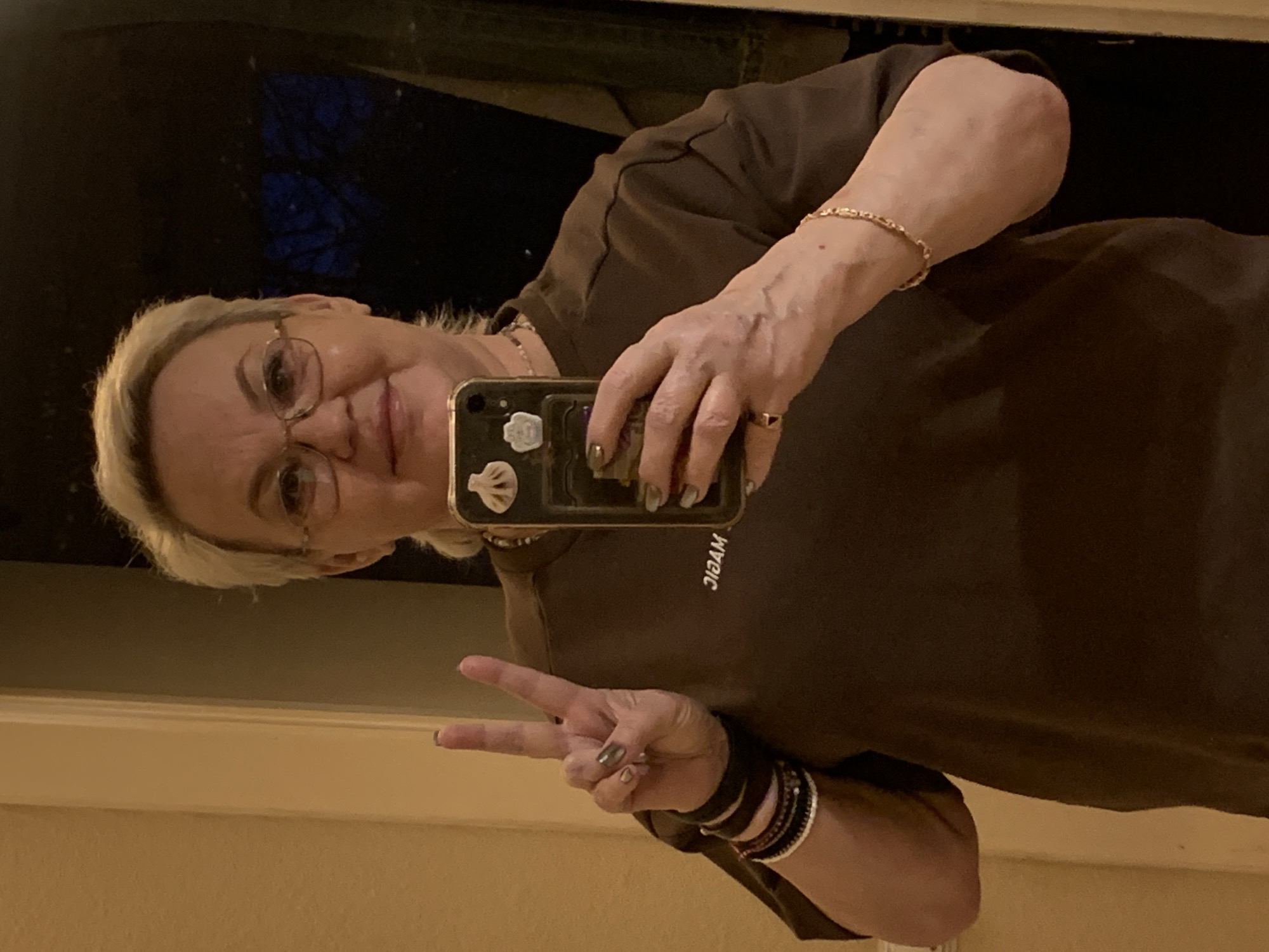 A woman with short blonde hair, wearing glasses and a brown T-shirt with the word "MAGIC" printed backward, is taking a selfie in what appears to be a mirror. She's holding her phone up and making a peace sign with her other hand. A bracelet is visible on her wrist, and the background suggests an indoor environment. The location is not discernible from this image.