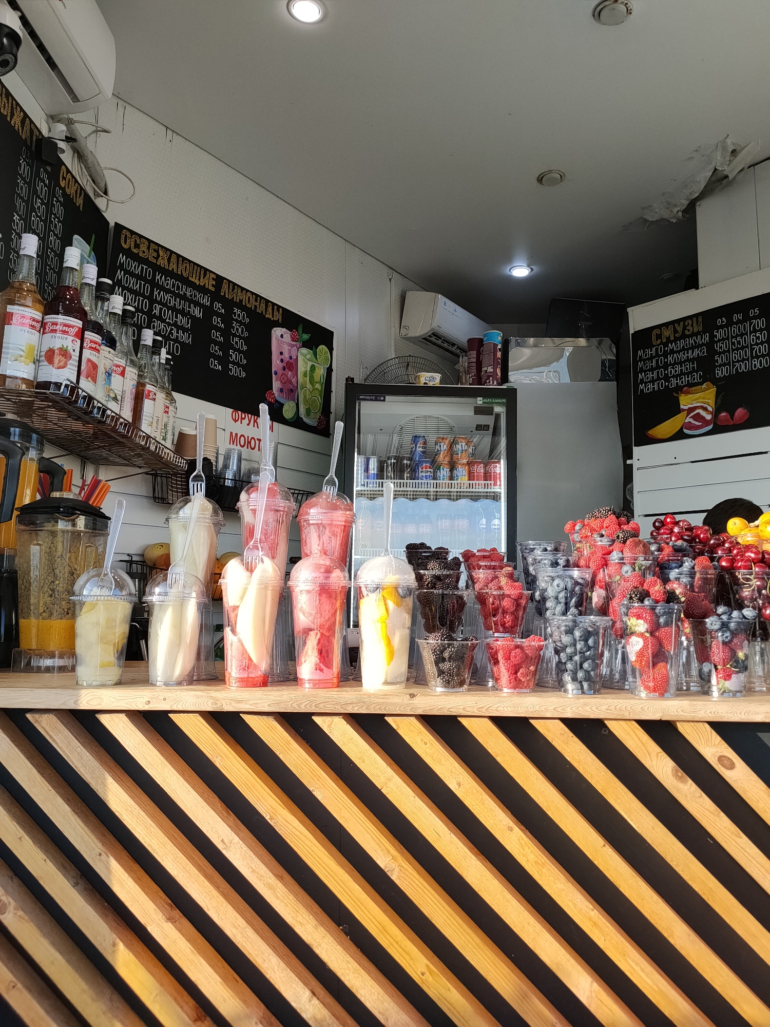 This is a medium shot, eye-level, outdoor, natural light photograph of a brightly lit smoothie and juice stand. The stand is constructed with a wooden slatted front, with the wood planks angled diagonally from lower left to upper right. Behind the counter, on shelves, are various bottles of syrups and juices, some with labels visible. To the right of the shelves, a black chalkboard menu is mounted on the wall, listing various drinks with prices. Behind the menu, a glass-doored refrigerator unit is visible, filled with canned beverages.
On the counter, a large blender is positioned to the left, partially filled with a yellow liquid. In front of the blender and extending across the counter are several clear plastic cups filled with fruit and smoothies. These include cups with what appear to be sliced yellow fruit (possibly mango or pineapple), creamy white smoothies, and pinkish-red smoothies with visible fruit pieces. To the right of these cups are rows of smaller clear plastic cups filled with various berries and fruits, including blackberries, raspberries, blueberries, and strawberries. A few larger fruits, like cherries and lemons, are also visible in containers.
In the background, the interior of the stand is visible, with white walls and some equipment. A large fan is mounted on the left side of the stand. The overall impression is of a busy, well-stocked food and beverage vendor, likely in a public area. The presence of Cyrillic script on the menus and beverage labels, along with the mention of Sochi, Russia in the prompt, strongly suggests the location.