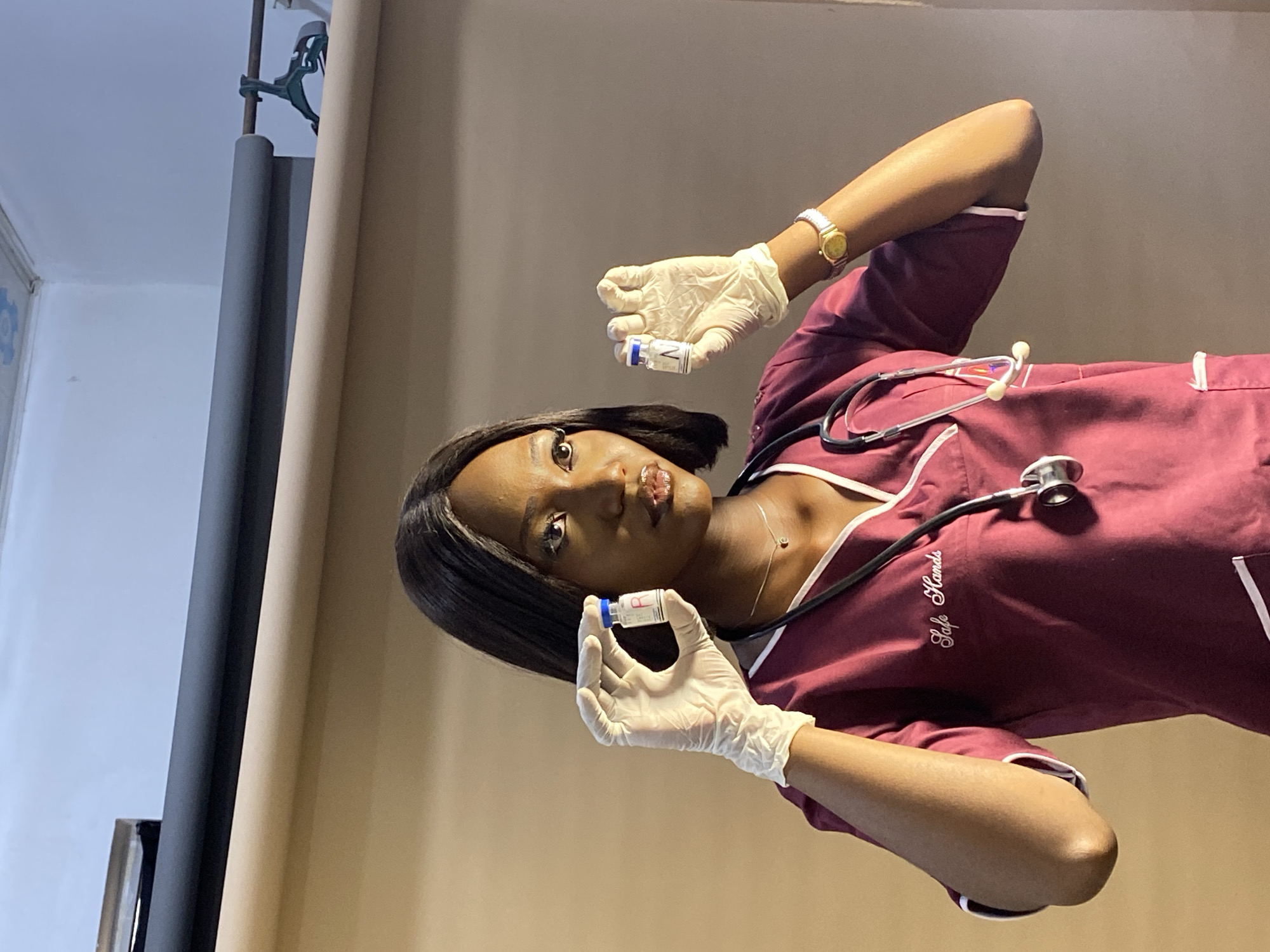The image shows a dark-skinned woman with short black hair wearing a maroon scrub top with white trim, a stethoscope around her neck, and white gloves. She is holding two small vials, one in each gloved hand. The background consists of a beige backdrop and a black stand. The location is presumably indoors, but there is no specific information in the image to identify it as being in Gura Top, Nigeria.