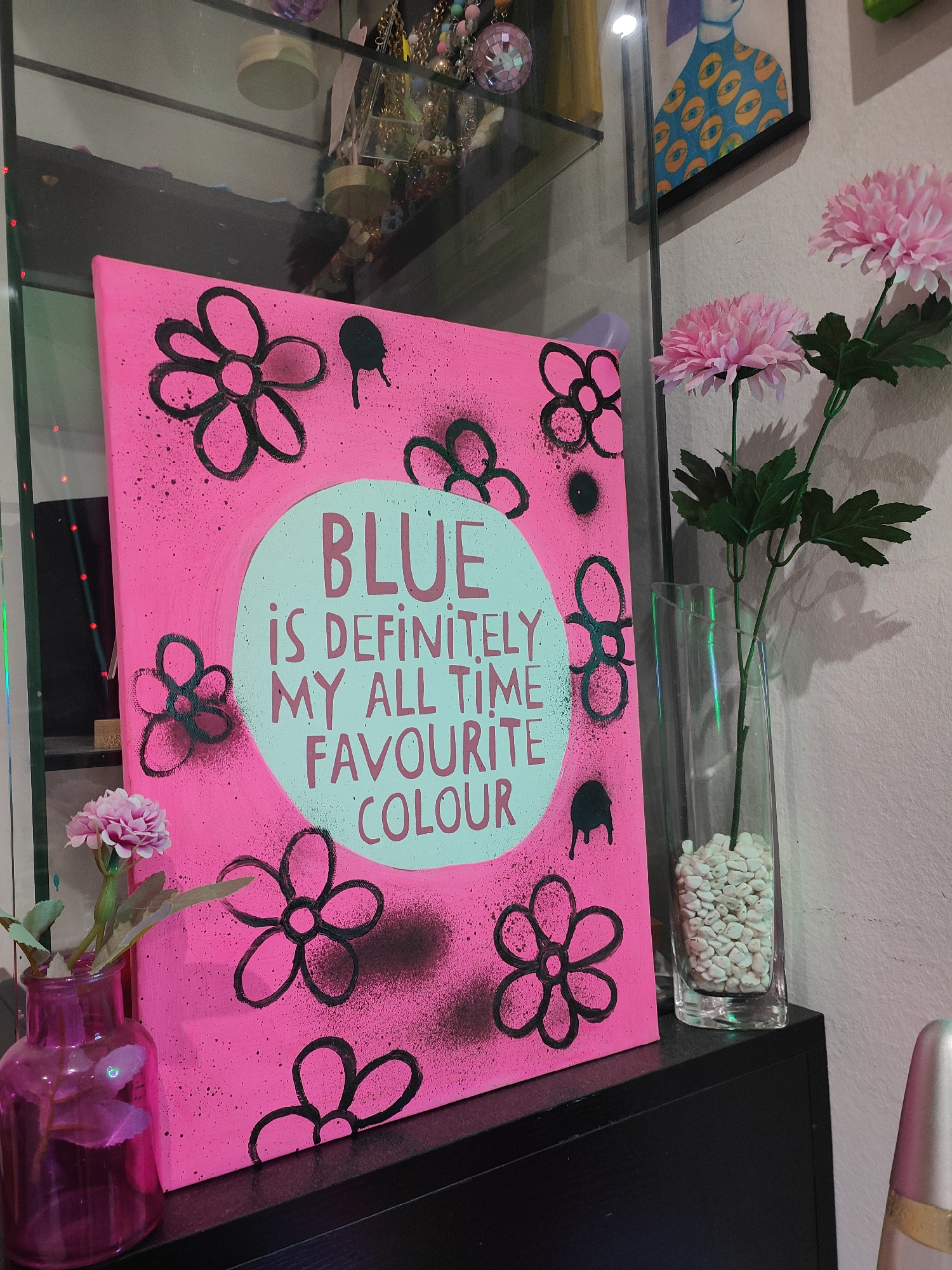 An indoor scene showcases a vibrant, neon pink canvas painting as its focal point, resting on a dark, flat surface, likely a shelf or cabinet. The painting features several black outlined flower motifs, accentuated by black spray paint splatters and drips around them. Centered on the canvas is a light blue oval containing the text, "BLUE IS DEFINITELY MY ALL TIME FAVOURITE COLOUR," written in capital letters.
To the right of the painting, two large artificial pink chrysanthemums with green stems are displayed in a tall, clear glass vase filled with white pebbles. To the left, a smaller single artificial pink flower is held in a small, dark pink translucent glass vase.
The background on the left reveals a glass display cabinet housing an array of colorful decorative objects, possibly jewelry. On the right, behind the flowers, a section of a light-colored wall is visible, with a framed artwork depicting a person with a blue, eye-patterned shirt hanging on it. The overall impression is one of a cheerful and artistic decorative display, likely within a home setting. There is no activity taking place.