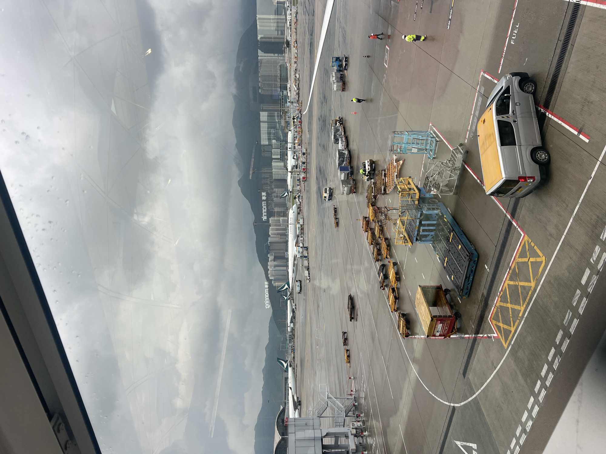 This image depicts a wide-angle view of an airport tarmac on a cloudy day, likely taken from an airplane window. The scene shows multiple parked aircraft with the distinct green and white livery of Cathay Pacific Airways. Several baggage carts, some loaded with cargo, are spread across the tarmac. Various airport service vehicles, including a silver van with a yellow roof and a small utility vehicle, are also visible. In the background, a densely populated urban area with high-rise buildings and a mountain range under a heavy, overcast sky can be seen, which is characteristic of Hong Kong. The tarmac appears wet, indicating recent rain. The overall impression is one of a busy but somewhat subdued airport environment due to the weather.
