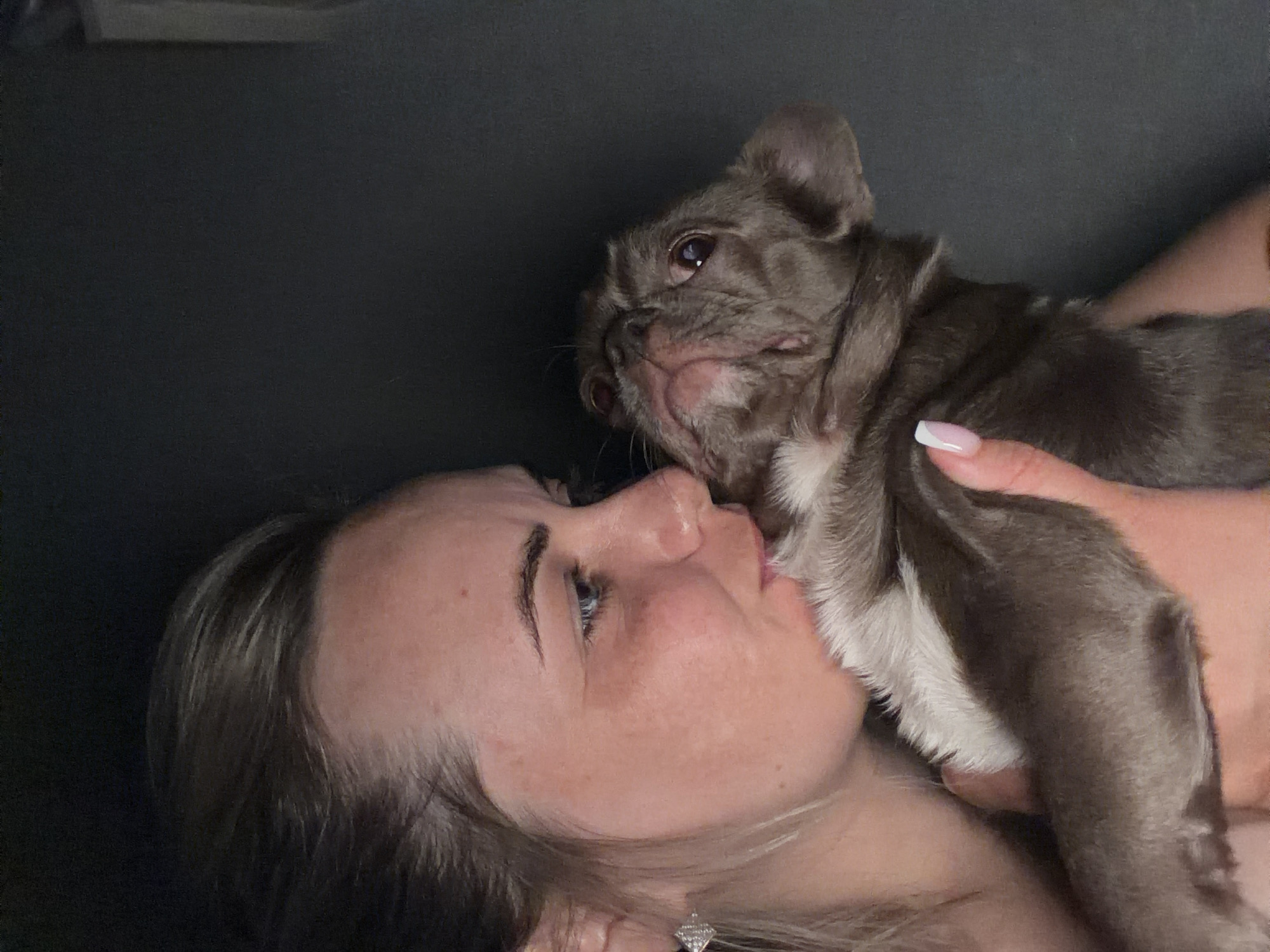 A woman lies on a dark surface, holding a gray and white French Bulldog close to her face. The dog is pressed against her cheek, and the woman looks upwards towards the dog. The woman has light skin, light brown hair, and blue eyes. She wears a small stud earring. Her nails are manicured with a French tip. The environment appears to be indoors. There is no indication of this image being in the city of Votkinsk, Russia.