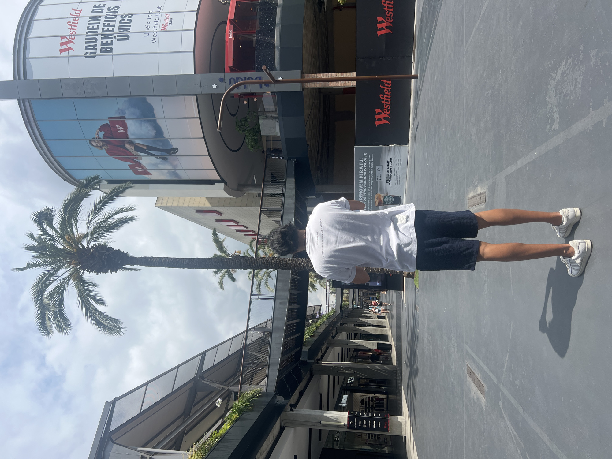 A person stands facing away from the camera in the middle of an outdoor walkway with shops on either side. The person is wearing a white t-shirt with text on the back and dark blue shorts. They are also wearing white sneakers. The walkway is paved with a grey material and lined with planters containing greenery. A tall palm tree is visible to the left of the person, and another is further in the background.
In the background, a large building with a curved glass facade displays advertisements. One advertisement features a woman sitting on a cloud, and another has the "Westfield" logo. The text on the advertisements is in Catalan. Signs for "Tezenis" and "Massimo Dutti" are visible on the storefronts.
The environment appears to be a modern outdoor shopping area, likely a mall or retail complex. The sky is partly cloudy. Based on the Catalan language on the advertisements, the location is the Westfield La Maquinista shopping center in Barcelona, Spain.