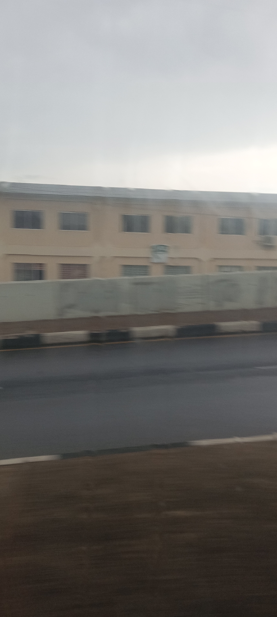 The image presents a view of a street scene in Jos, Nigeria, characterized by an overcast sky. A beige, two-story building stands behind a concrete barrier. Windows are visible on both floors. An air conditioning unit is mounted on the building. A road runs in front of the barrier, showing signs of wetness, possibly from rain. The foreground appears to be a roadside area.
