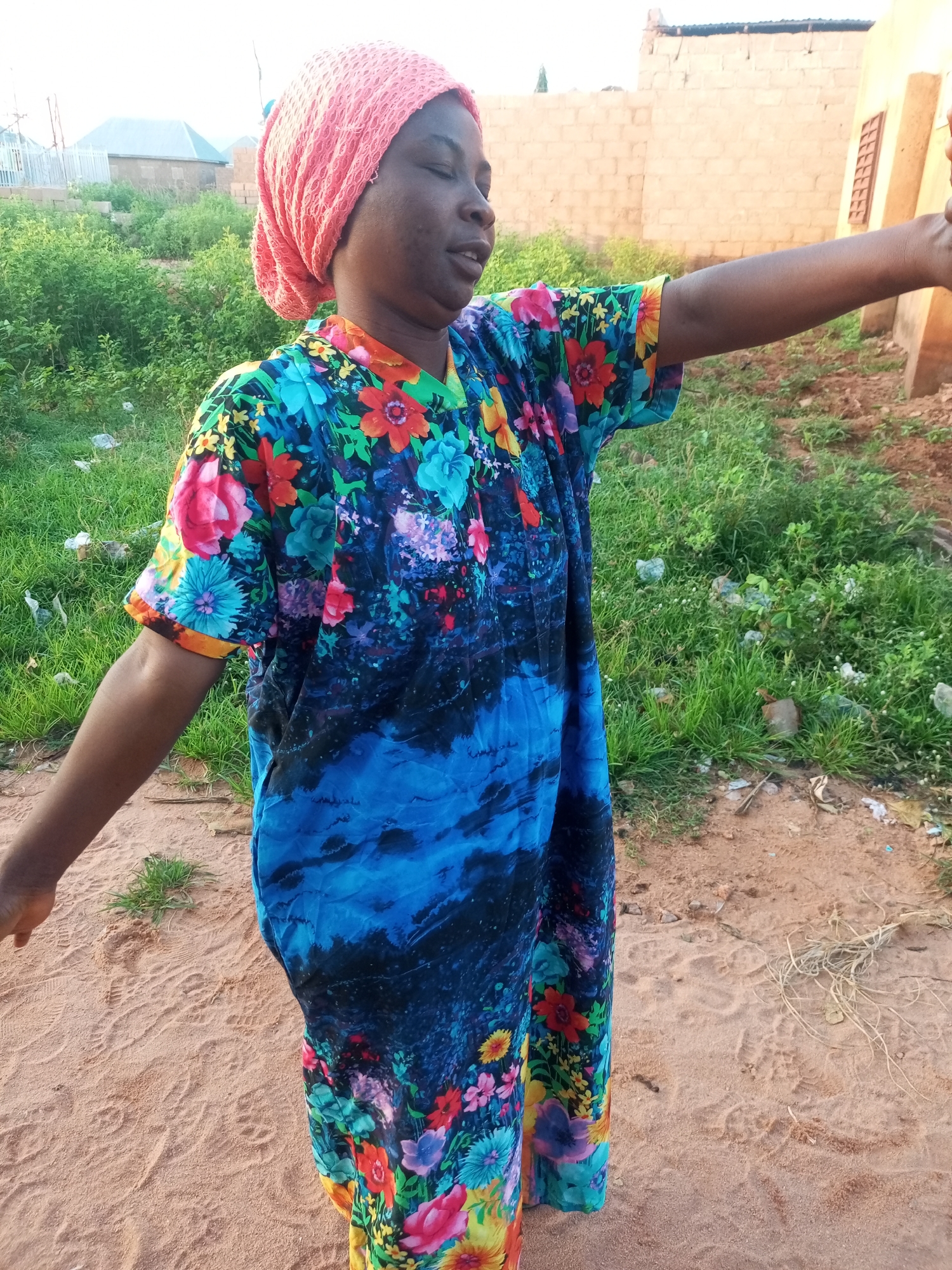 A full-body, vertical shot shows a Black woman from the knees up, standing in a sandy, outdoor area. She is wearing a bright, floral-patterned dress with blue, pink, yellow, and green flowers and a pink headscarf. Her right arm is extended, with her hand out of frame, as if she's reaching for something or waving. The background consists of green foliage, dirt, and buildings constructed of light-colored brick. The scene is set in Tempure, Nigeria.