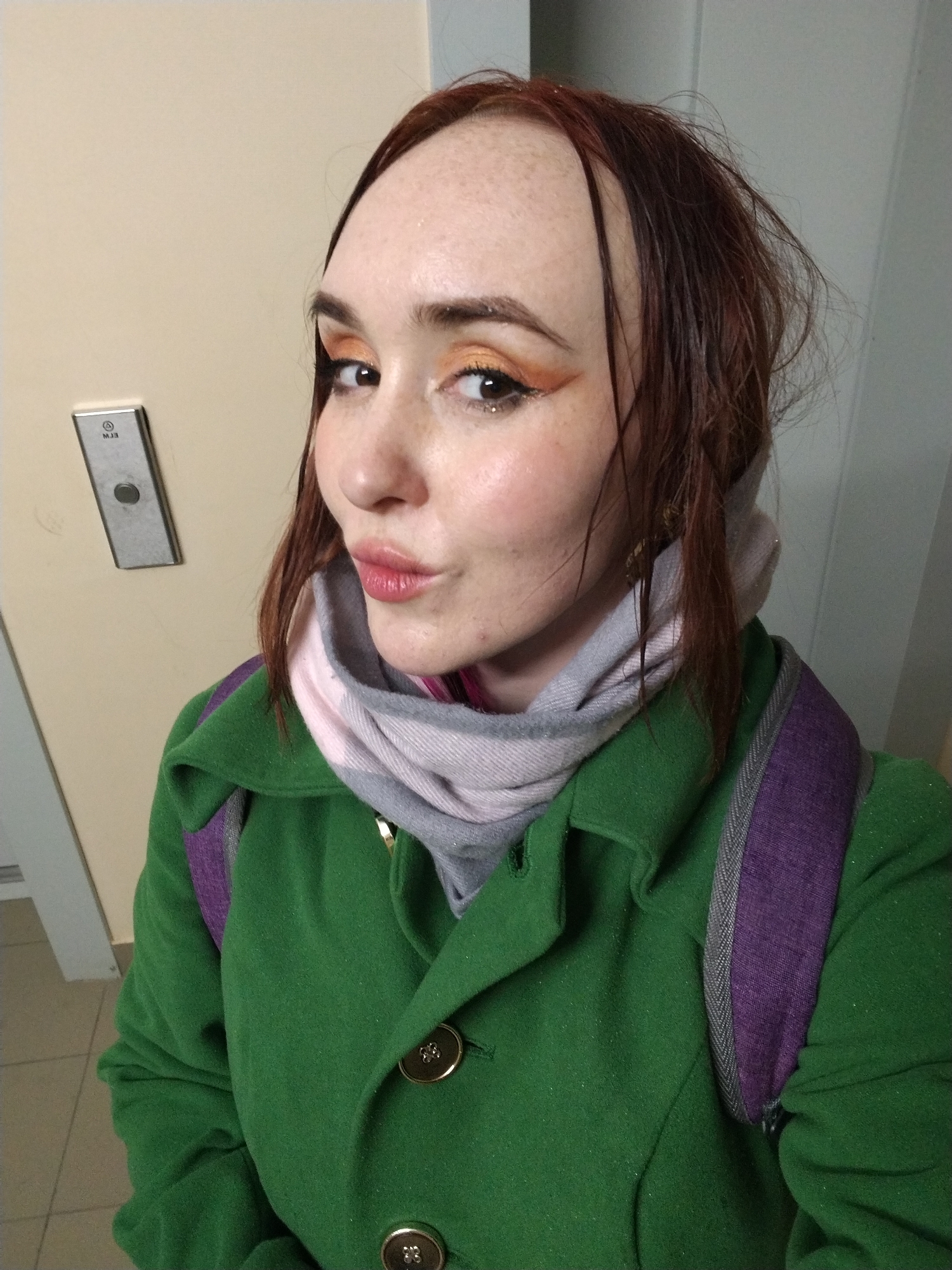 A selfie-style portrait captures a young woman indoors, likely in a building hallway or corridor in Izhevsk, Russia. The woman, with reddish-brown hair, freckles, and a playful expression featuring puckered lips, looks directly at the camera. She wears vibrant makeup, including orange eyeshadow and pinkish-red lipstick. Her attire consists of a dark green coat with large gold buttons and a gray and light pink plaid scarf wrapped around her neck. A purple backpack strap is visible over her right shoulder.
To the left of the woman, mounted on a light beige wall, is a silver rectangular panel with a central circular button, characteristic of an elevator call button. Upside down on this panel, faintly visible text resembling "MЭЗ" is present, which could be a Cyrillic label or part of a floor indicator, consistent with the Russian location. The floor is light-colored and tiled. The overall scene suggests she might be waiting for an elevator or paused in a hallway. Her warm clothing indicates cool weather conditions, which aligns with the climate in Izhevsk.