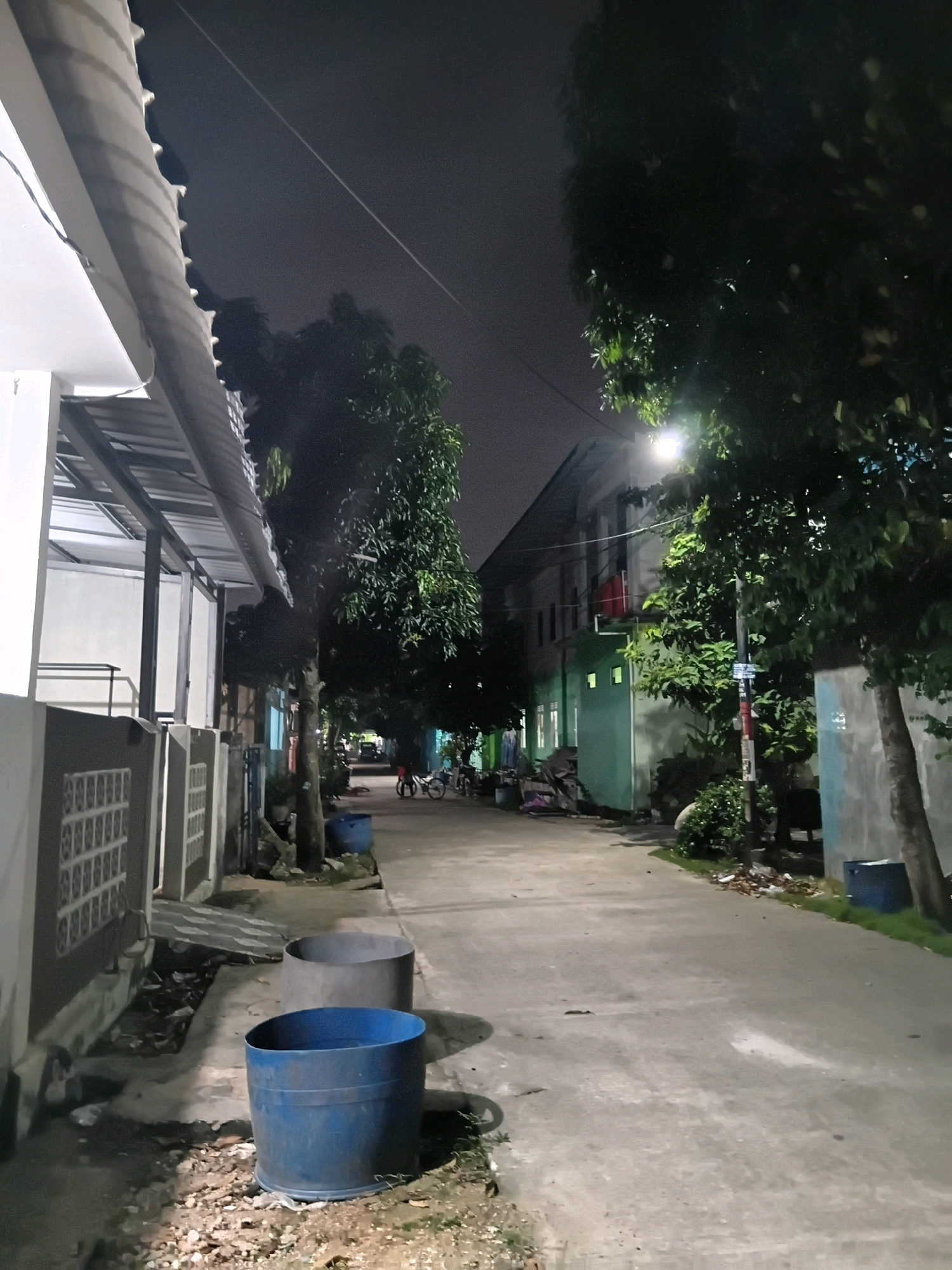 An outdoor, low-angle, nighttime shot shows a concrete alleyway between buildings. On the left, a white building with a grey, corrugated metal awning extends into the foreground. A grey concrete wall with a decorative, repeating pattern runs along the front of the building. In the center of the image, the alleyway stretches into the distance, illuminated by a streetlamp. On the right side of the alley, a green, two-story building has windows and an open doorway. Trees with lush green foliage line both sides of the alleyway. In the foreground, two large blue and grey cylindrical bins sit on the side of the alley. Further down the alley, a few people can be seen walking or standing near parked bicycles. The sky is dark, with a faint white line visible in the upper portion of the image, possibly a contrail. The overall scene depicts a residential or semi-residential area at night.