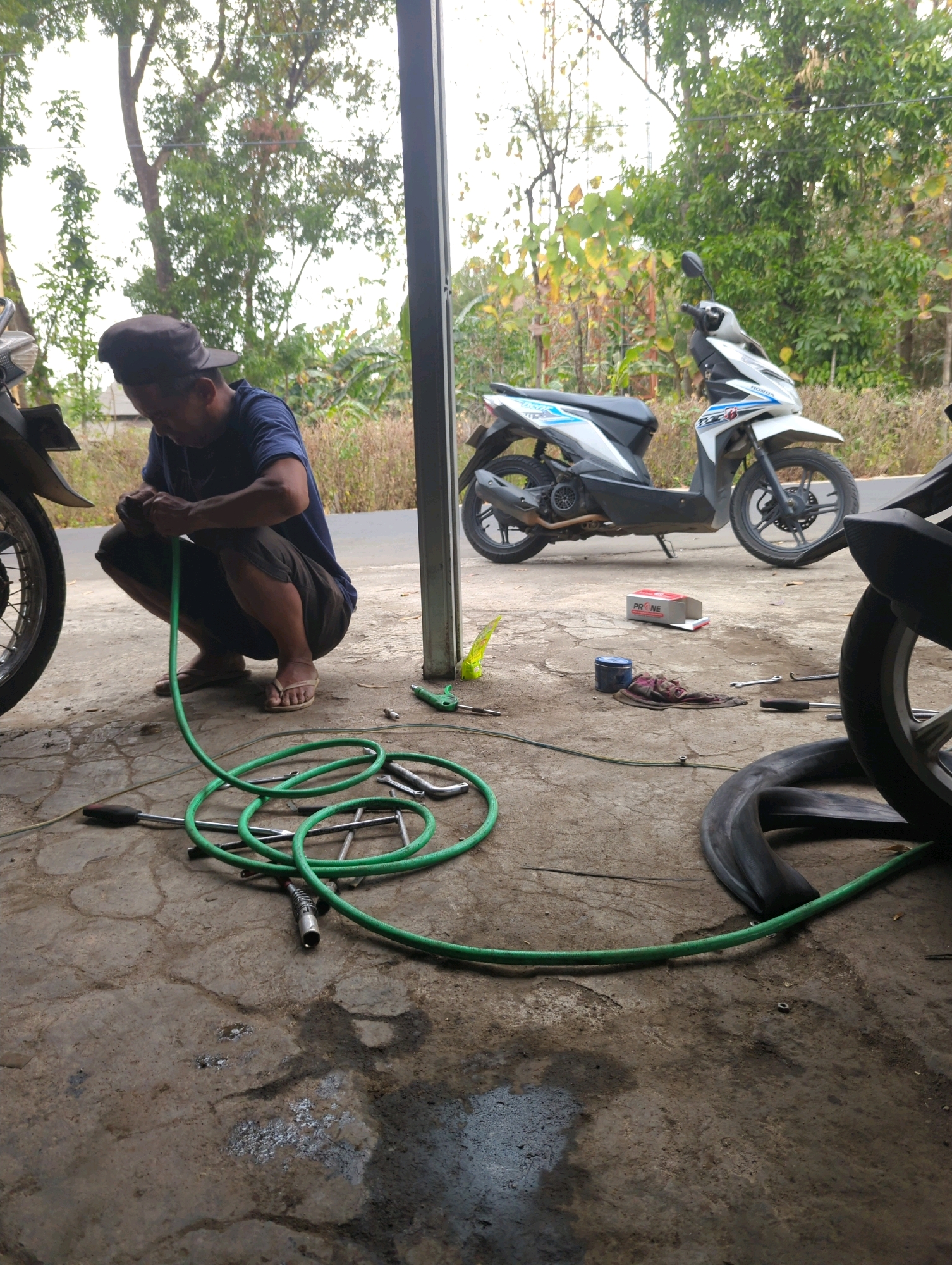In Kedungdowo, Indonesia, a person is squatting on a concrete surface, working on a green hose. The person wears a dark blue t-shirt, brown shorts, a dark baseball cap, and flip-flops. The hose is coiled on the ground, with one end near the person's hands and the other extending to the right. Various tools are scattered around, including wrenches, a screwdriver, and an air nozzle. In the background, two motorcycles are parked. One is a white and blue scooter, and another is partially visible on the left, appearing to be a dark-colored motorcycle. The environment suggests an outdoor workshop or repair area, with trees and foliage visible in the distance. The ground is a textured, cracked concrete with some patches of dirt and moisture.