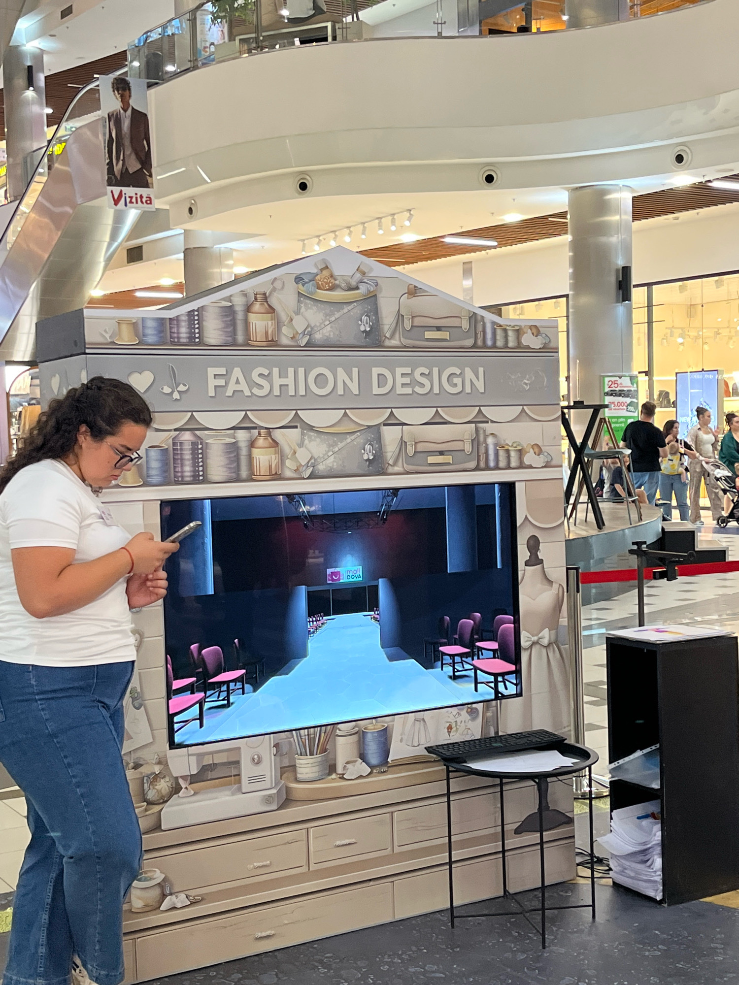 The image shows a woman standing in a shopping mall, looking at her phone. She is wearing a white t-shirt and blue jeans. In front of her is a large display featuring the words "FASHION DESIGN". The display has a screen showing a runway with pink chairs. To the right of the display is a black shelf unit filled with papers. In the background, other shoppers are visible, as well as escalators and storefronts. The setting appears to be an indoor shopping mall. There is no indication of the city of Chișinău, Moldova in the image or its accompanying crops.