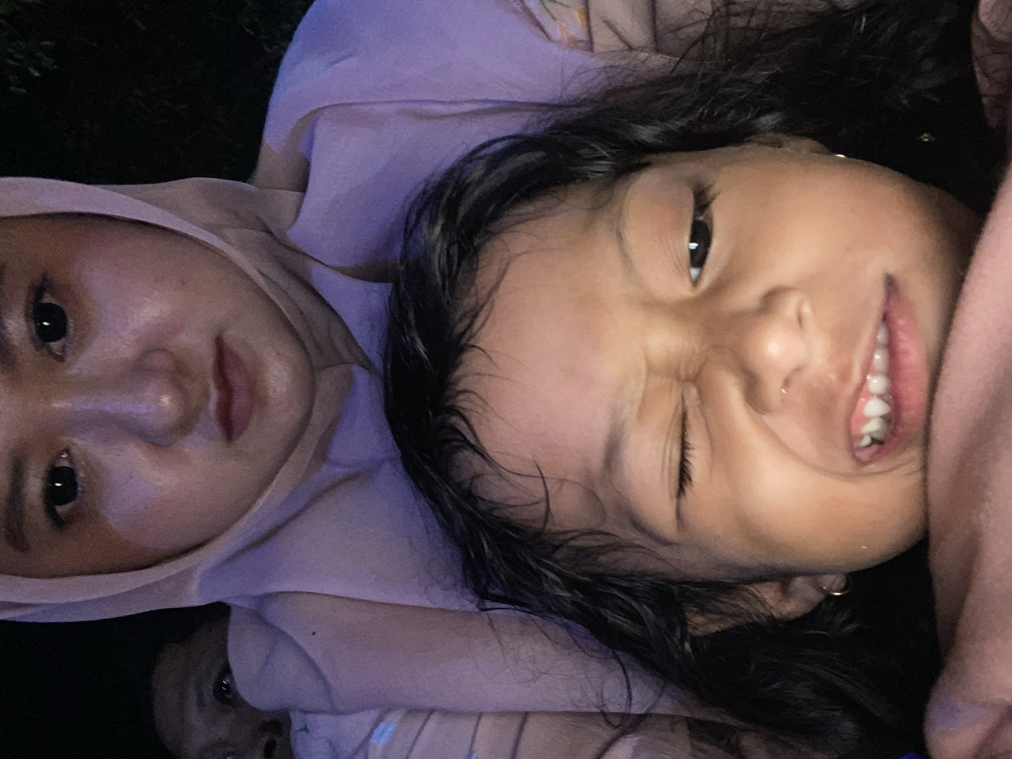 In Sidoarjo, Indonesia, a close-up, low-angle selfie features a young child in the foreground, her face contorted in a grimace. She is winking with her right eye, and her mouth is open, revealing her teeth. Her dark, curly hair frames her face, and she is wearing a small gold hoop earring. Behind her, an adult wearing a pink hijab and matching garment is visible, their expression neutral or slightly stern. Another person's face is partially visible in the dark background on the left. The lighting is dim, with a warm, purplish hue illuminating the scene.
