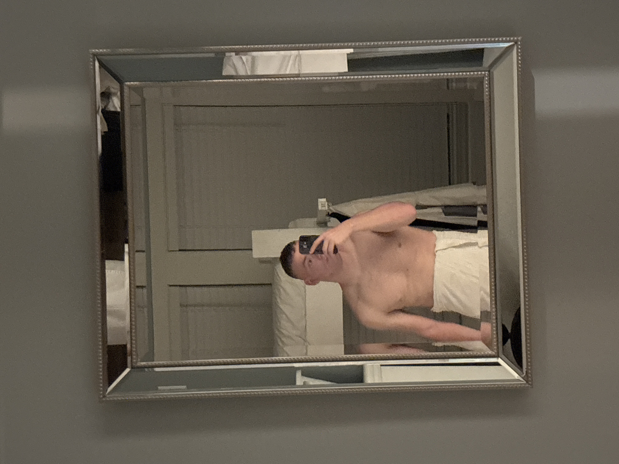 The image shows a person taking a selfie in a mirror. The person is shirtless and wearing white shorts or a towel. The background appears to be a bedroom with a bed and some furniture. The location is Panama City Beach, United States of America.