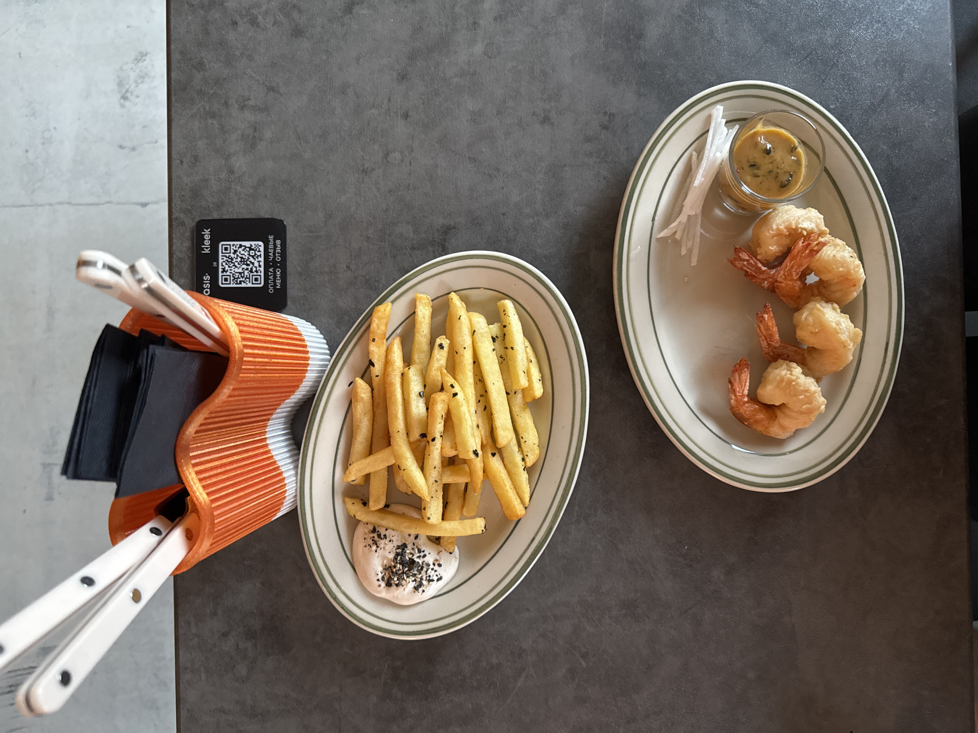 An overhead shot shows two plates of food on a dark gray, textured surface. The plate in the foreground contains four breaded shrimp, a small glass of sauce, and thinly sliced white radish. The plate behind it holds a portion of french fries with a small dish of white sauce sprinkled with black sesame seeds. To the left of the plates, a white and orange utensil holder contains napkins and cutlery. A black QR code sticker is visible on the table next to the utensil holder. The sticker has the text "asis kleek" and a QR code. Below the QR code, there is text in Russian which appears to be related to payment, tips, menu, and reviews. The lighting suggests an indoor setting, possibly a restaurant.