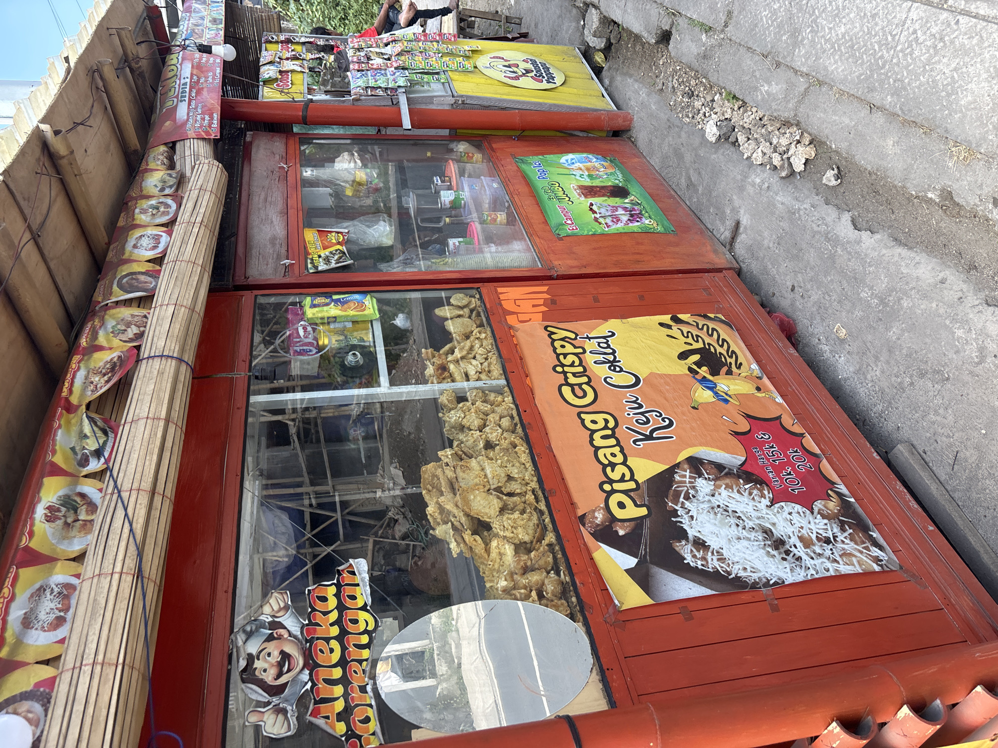 The image displays a series of vibrant Indonesian street food stalls, predominantly red and yellow, set up alongside a paved sidewalk in Kuta Selatan, Indonesia, under bright daylight. The stalls are oriented vertically in the frame, suggesting the photo was taken with the camera tilted.
The main subjects are the various food items and their advertisements. Several glass display cases showcase an assortment of fried snacks, known as *gorengan*, including what appears to be tempeh, a popular Indonesian staple. Packaged snacks and drinks are also visible within the displays.
Prominently featured banners and signs advertise the offerings. One large orange banner on the front of a stall promotes "Pisang Crispy Keju Coklat" (Crispy Banana Cheese Chocolate), showing a picture of the dessert topped with grated cheese and chocolate sauce. Pricing options of "10k, 15k & 20k" (10,000, 15,000, and 20,000 Indonesian Rupiah) are listed. Another sign with a cartoon chef mascot reads "Aneka Gorengan" (Various Fried Snacks). A red banner above details items "SEDIA:" (Available:), listing "Pisang Keju Susu Coklat," "Tape," "Pisang Goreng," "Tempe," and "Bakwan." Further down, a green sign advertises "Es Campur JUMBO Pop Ice" (Jumbo Mixed Ice Pop Ice). A yellow cart in the background features the text "Sosis Halal."
The scene depicts a typical outdoor food vendor setup, ready to serve customers. In the background, a person's legs are visible near the yellow cart, suggesting a vendor or customer. The general ambiance is bustling and casual, characteristic of street food culture. A roll-up bamboo blind is seen on the left side of the stalls, possibly used for shade or closing the stall. The ground is paved concrete with a narrow ditch containing small rocks.