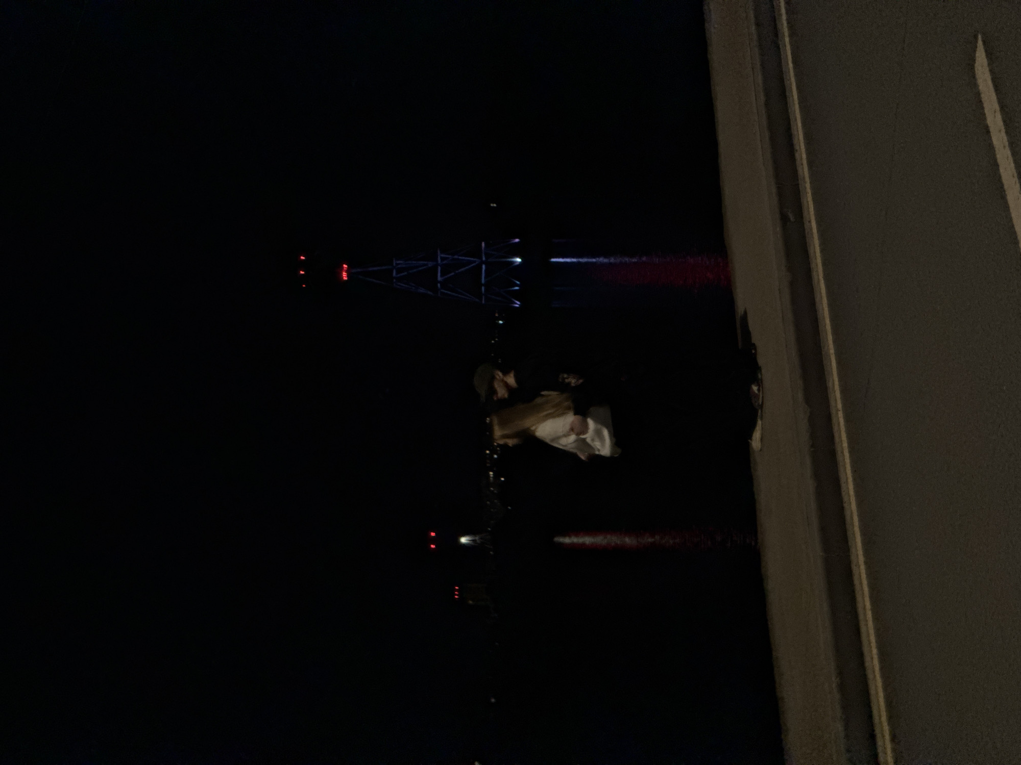 At night in Nizhny Novgorod, Russia, a woman stands on a concrete structure and faces a dark body of water with reflections of lights. A large structure is illuminated with blue and red lights across the water. The woman has long blonde hair and is wearing a dark hat and light-colored clothing.