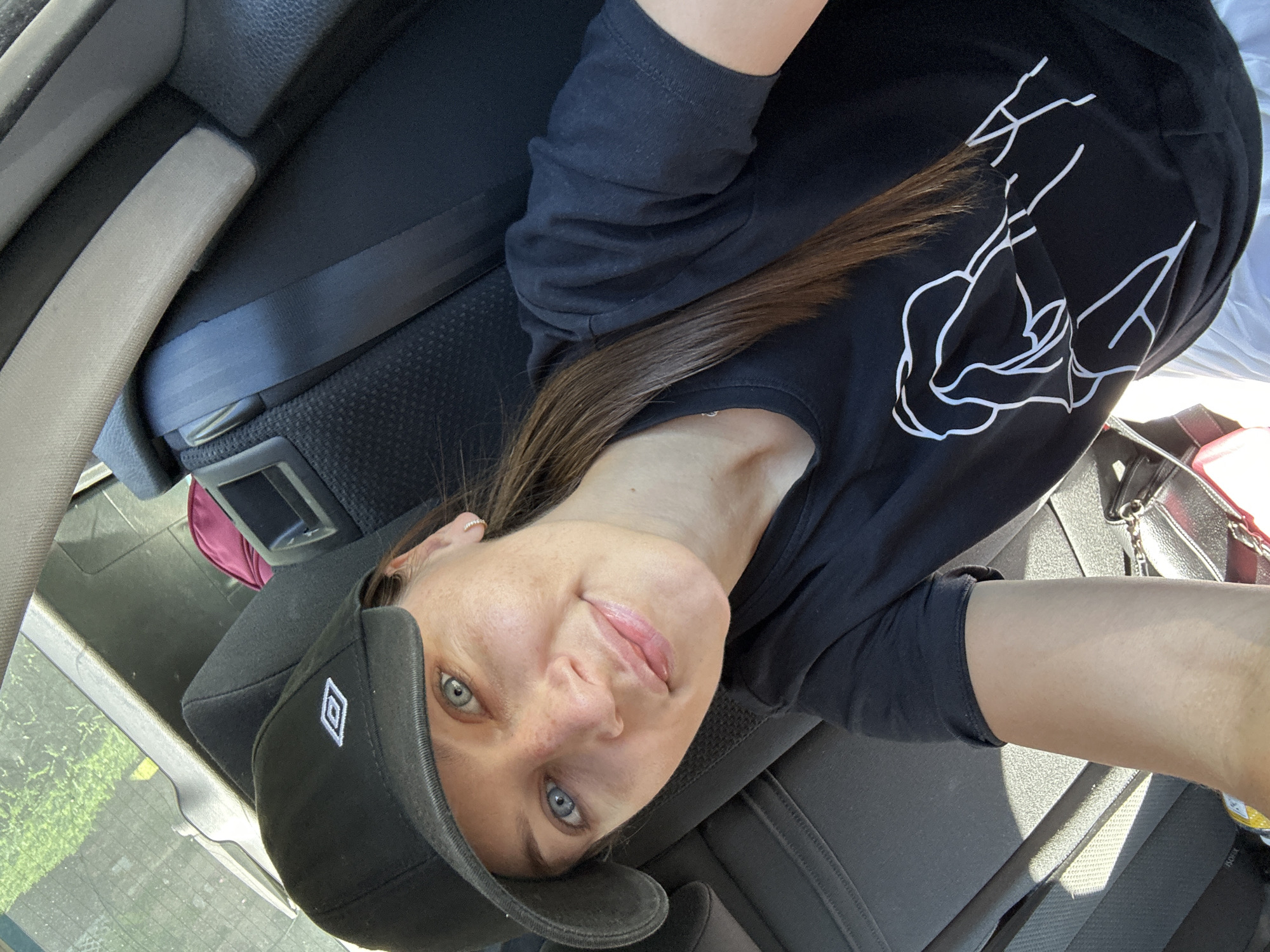 A young woman with fair skin and blue eyes is seated in a vehicle. She wears a black baseball cap with a white logo and a long-sleeved black shirt with a white graphic design. The vehicle's interior is visible, including a seatbelt and seat. The photo appears to be taken from a first-person perspective, looking slightly downward. The location is possibly near Niederquembach, Germany, based on the analysis request.
