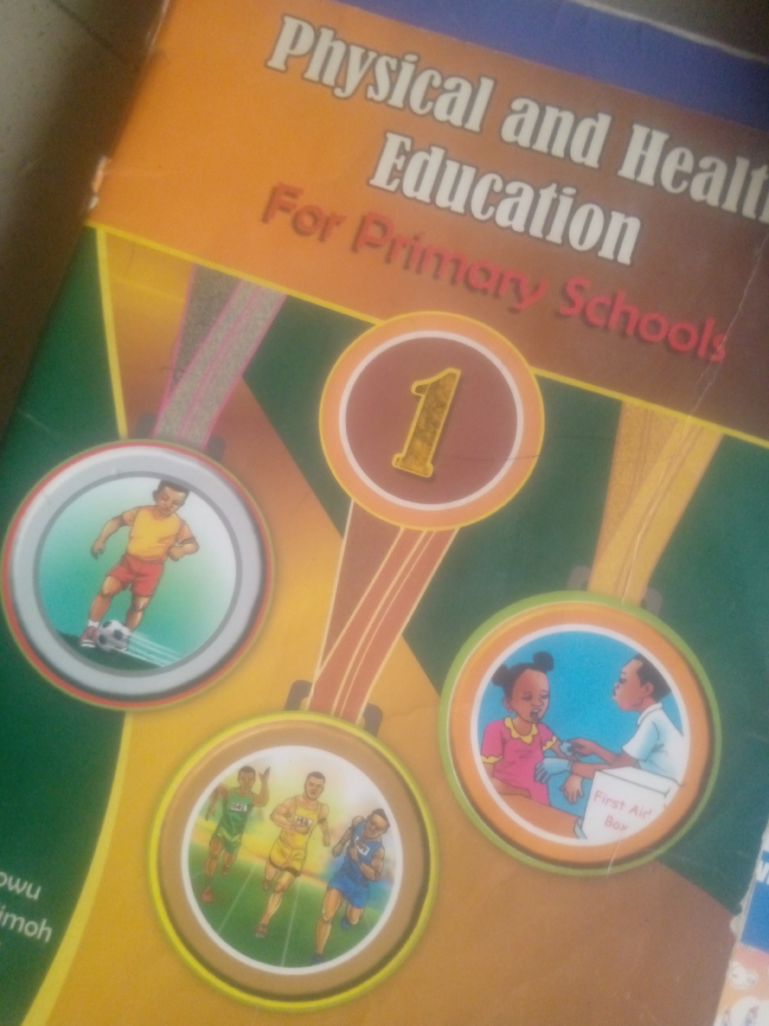The image is a close-up, top-down shot of the cover of a book titled "Physical and Health Education For Primary Schools". The cover features an orange and blue background with graphics of medals. One medal depicts a boy playing soccer, another shows three runners, and a third illustrates a girl receiving first aid from a boy. A large number "1" is also prominently displayed in a medal-like circle. The text on the book is white and red. The bottom left of the image shows partial text from what appears to be author names, "wuw" and "imoh". The overall lighting is dim, and the image quality is moderate, suggesting it was taken with a handheld device. No specific location in a city is identifiable from the image itself.