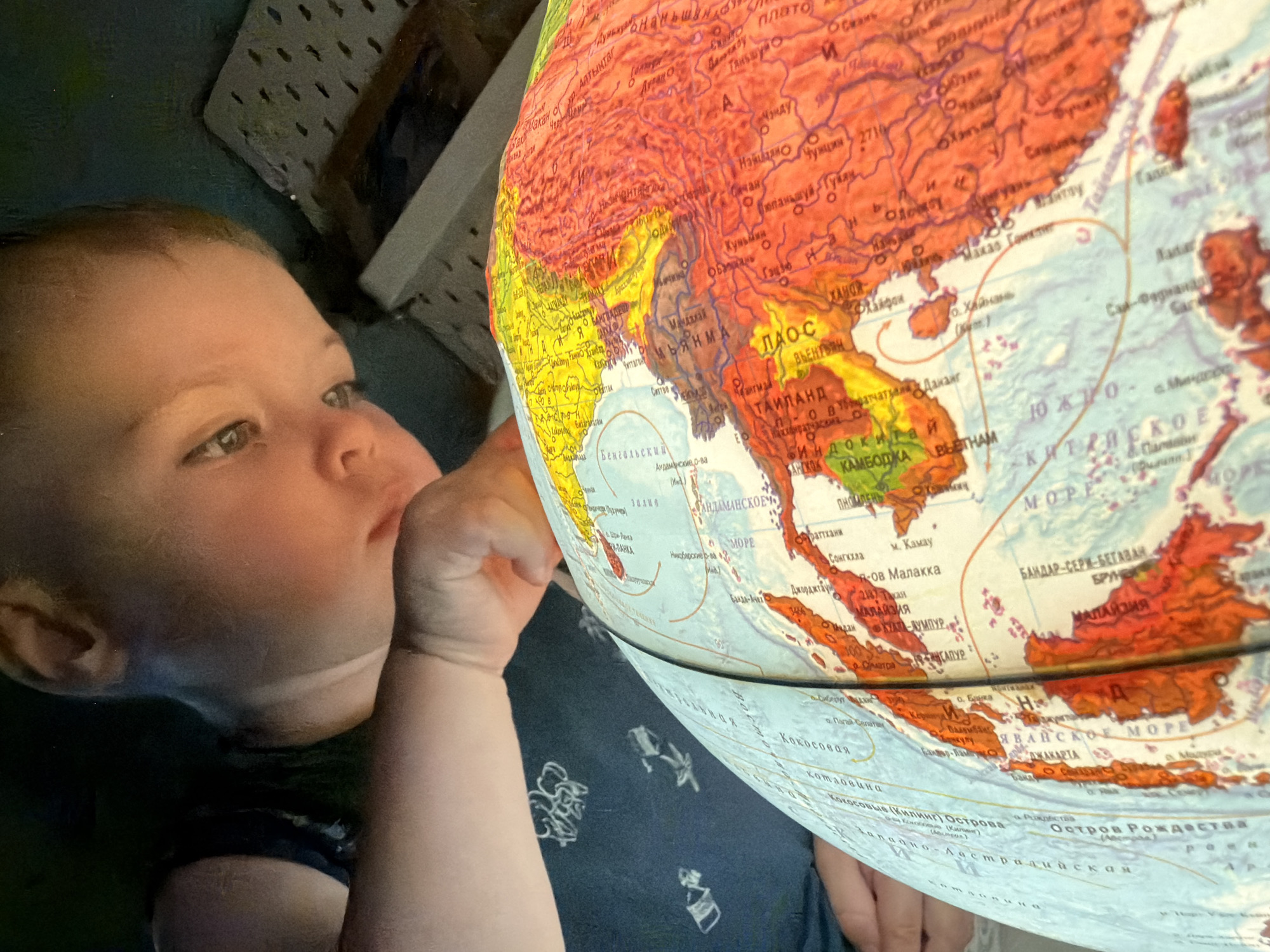 In the dimly lit setting of Votkinsk, Russia, a young child is engrossed in exploring a glowing terrestrial globe. The child, positioned on the left side of the frame, leans towards the globe, their face illuminated by its light, with their finger gently touching the surface. The globe displays various continents and countries in vibrant colors, with clear geographical features and text labeling locations in Russian. Prominently visible are parts of Asia, including Southeast Asia with labels such as "ТАЙЛАНД" (Thailand), "КАМБОДЖА" (Cambodia), and "ВЬЕТНАМ" (Vietnam). The child's focused expression suggests a moment of intense curiosity and discovery. The background is dark, with hints of furniture or shelving, creating an intimate and focused scene on the interaction between the child and the globe. The overall impression is one of quiet exploration and learning.
