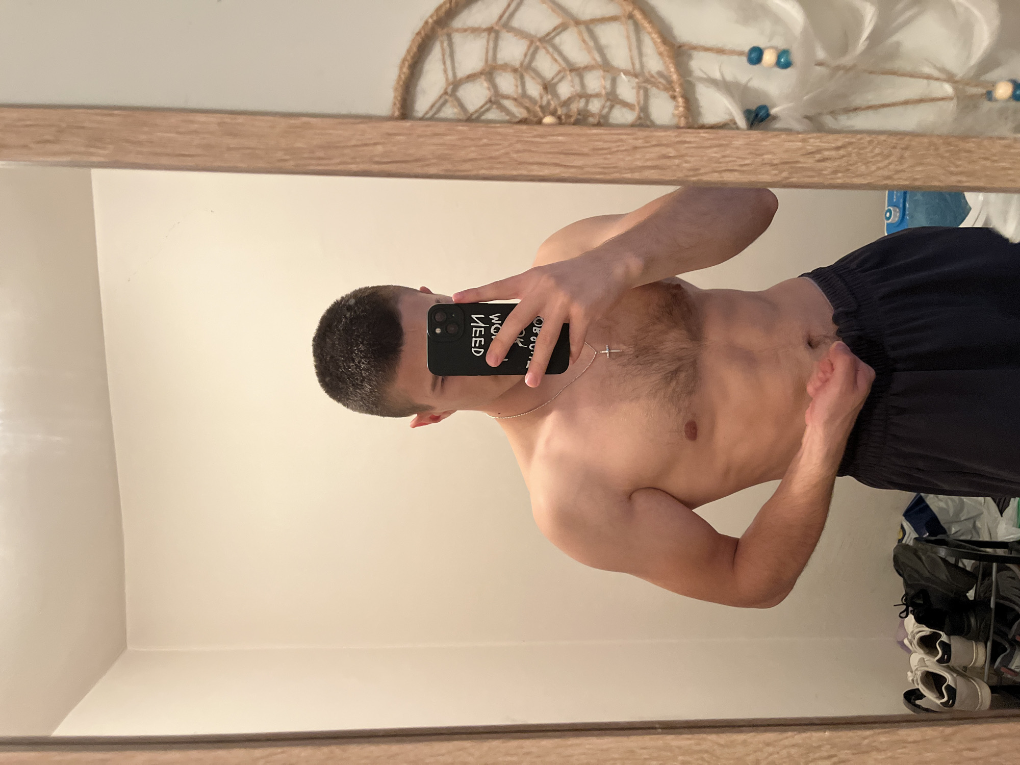 A shirtless man takes a mirror selfie, captured from a slightly elevated angle. He holds a black smartphone up to his face, obscuring part of his head. His dark hair is cut short. He has visible chest hair and wears a thin silver chain with a cross pendant around his neck. His body appears toned, and he is subtly flexing his left arm, with his right hand resting near his waist. He is wearing dark, possibly black or navy blue, elastic-waist shorts.
Visible text on the phone case, reflected in the mirror, appears as "WEED IW MON," which, when read correctly, is "I NEED NOW."
The background, reflected in the mirror, shows a light-colored wall. Above the man's head is a decorative, circular object resembling a dreamcatcher, made of woven rope. To the right of the dreamcatcher, there's a glimpse of a white feathery decoration with blue and white beads. In the bottom right corner of the reflection, a shoe rack or shelf holds several pairs of shoes, including white and black sneakers. A blue object, possibly a container or bottle, and a crumpled bag are also visible near the shoes. The overall lighting suggests an indoor setting.