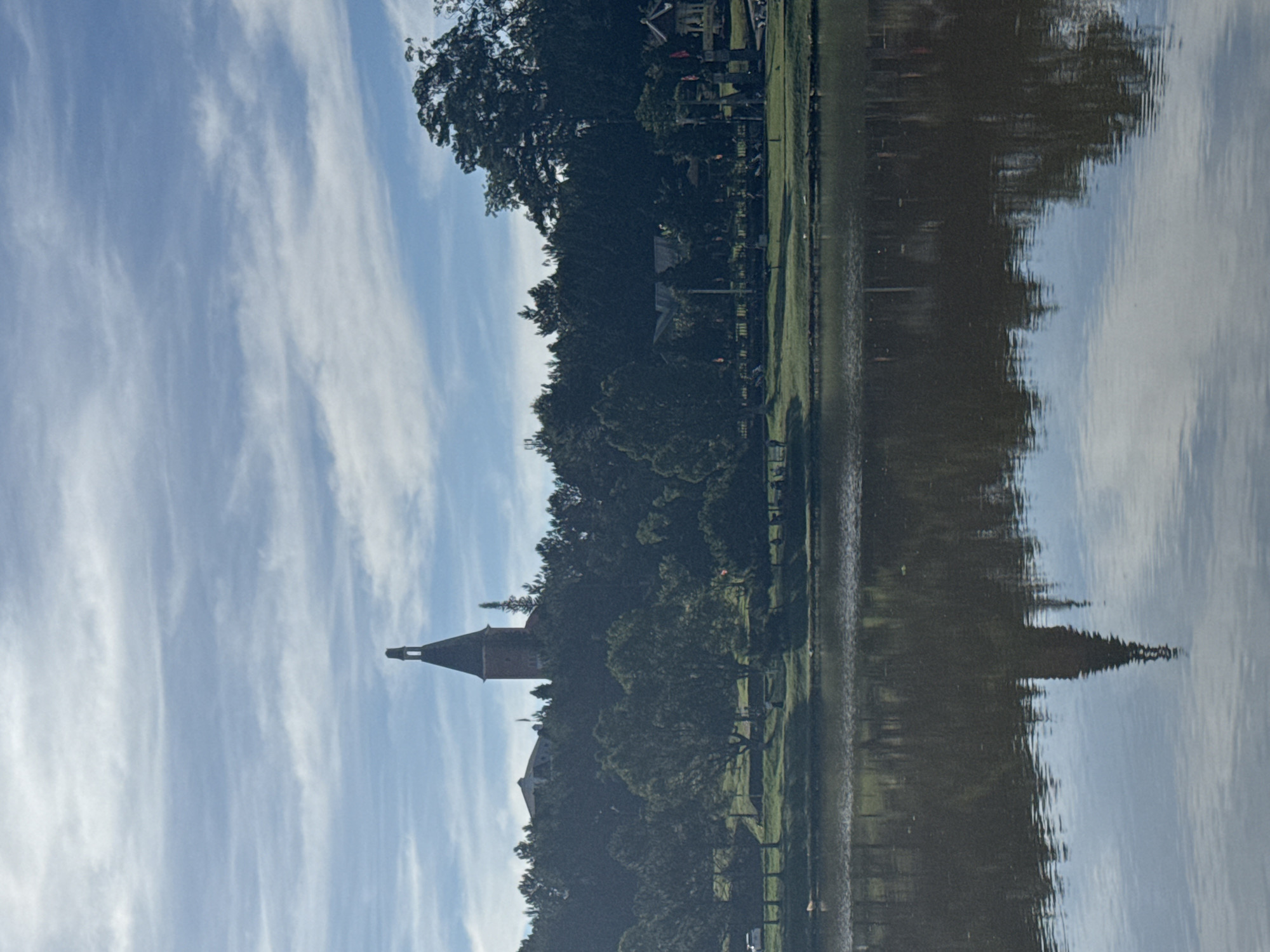 The image shows a body of water reflecting a building with a prominent spire, trees, grass, and a partially cloudy sky. The architecture suggests a European style. Given the city's European architectural influence and geographical features, it resembles Xuan Huong Lake in Da Lat, Vietnam. The reflection in the water is slightly distorted, indicating a slightly rippled surface. There's a treeline along the bank, and some structures are visible amidst the trees. The spire is the most prominent feature.