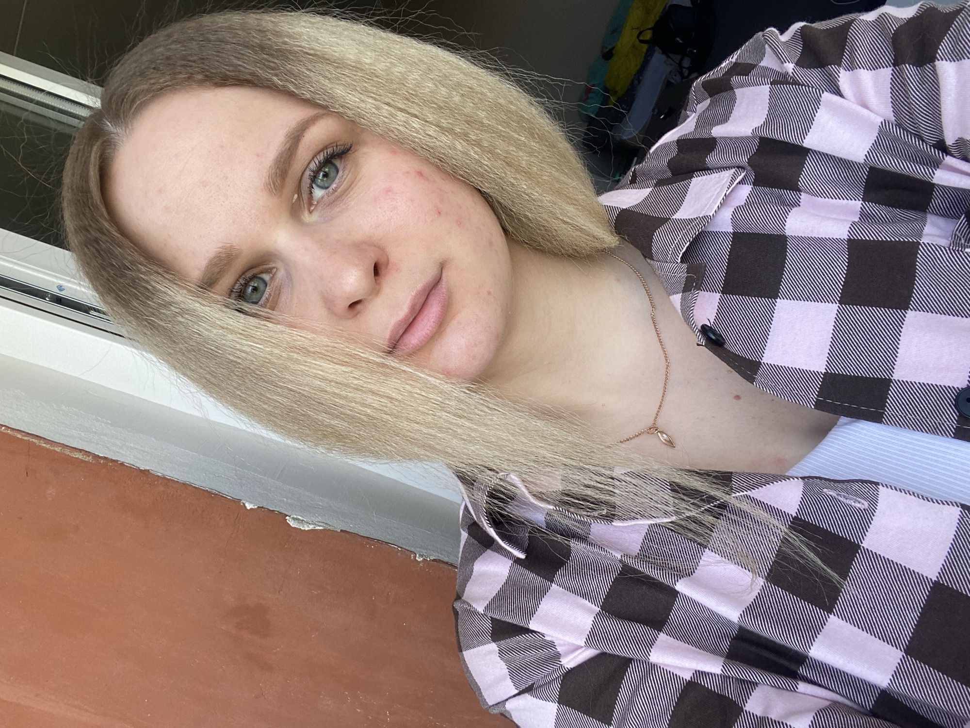 A selfie captures a young woman, likely located in Chaikovsky, Russia, looking directly at the camera with a neutral to slightly soft expression. She has long, blonde hair, which appears to have a slight crimped or wavy texture, partially framing her face. Her eyes are a light blue-green hue, and her lips are closed in a subtle, faint smile. Her fair skin shows some visible redness and minor blemishes on her cheeks and forehead.
She is dressed in a collared, button-up shirt featuring a distinctive light pink and dark brown/black plaid pattern. A plain white or light blue undergarment is visible beneath her plaid shirt. Around her neck, she wears a delicate gold-toned necklace adorned with a small, leaf-shaped or pointed pendant.
The background suggests an indoor or balcony setting. A white window frame is partially visible behind her left shoulder, and a reddish-brown textured surface occupies the lower left portion of the image, possibly a wall or railing. The area behind her right shoulder is darker and somewhat indistinct, indicating other objects or a cluttered space. The lighting suggests it is daytime. No visible text is present in the image.