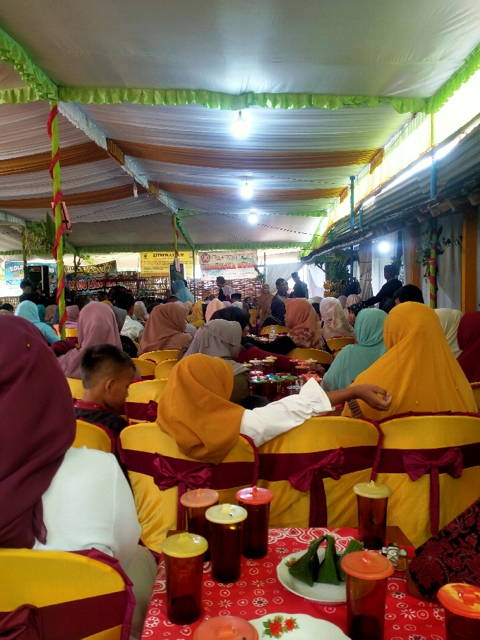 An event takes place under a tent with rows of people seated on yellow chairs decorated with burgundy bows. The majority of attendees are women wearing headscarves in various colors. Tables covered with red patterned cloths hold drinks in tall glasses with lids and small plates of food. The tent's ceiling is adorned with white and gold fabric and green trim, lit by hanging light bulbs. Banners and posters are visible at the front of the room.