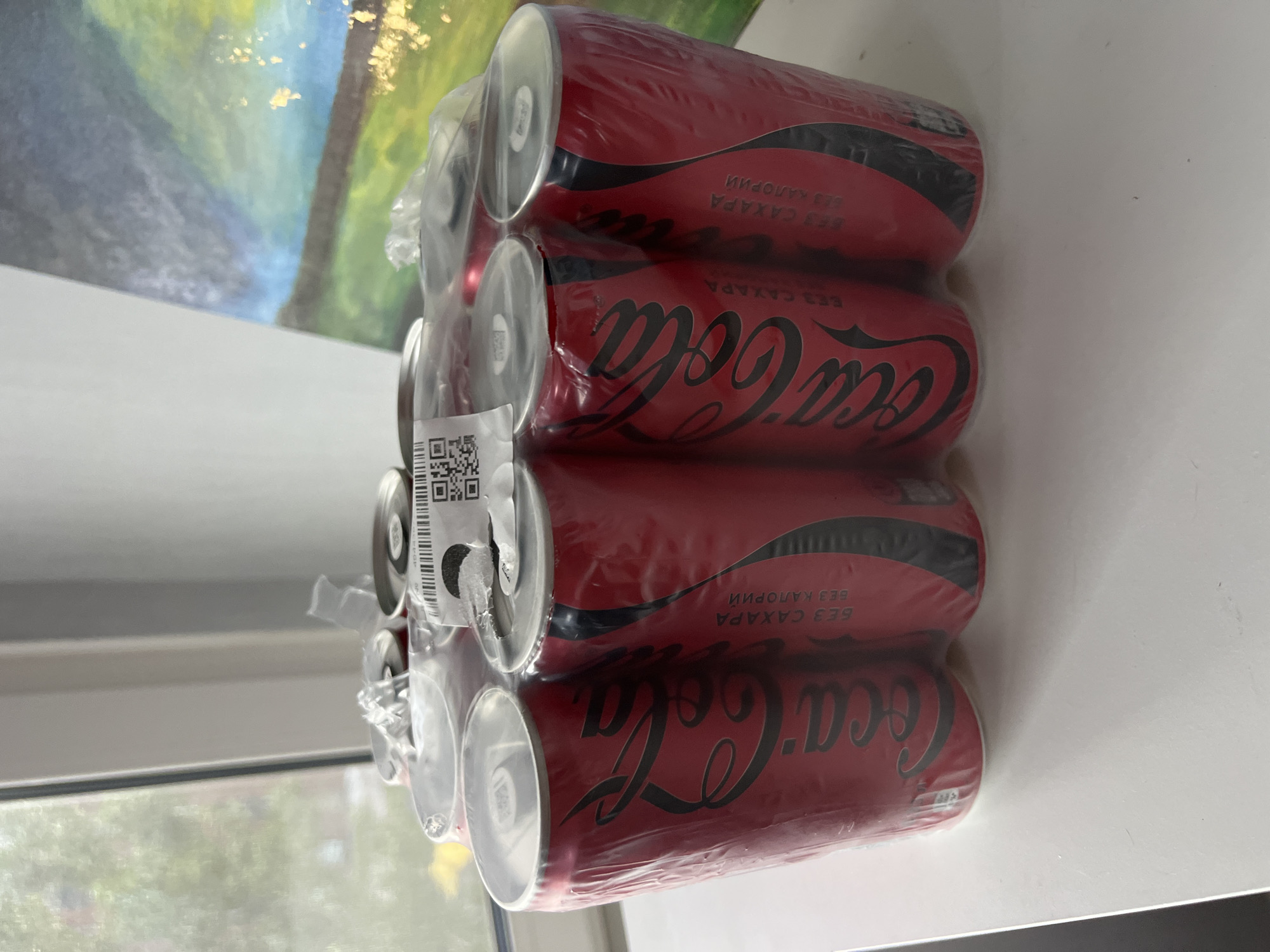 The image shows a six-pack of Coca-Cola Zero Sugar cans, wrapped in clear plastic. The cans are red with the Coca-Cola logo in black. Text on the cans reads "БЕЗ САХАРА" (no sugar) and "БЕЗ КАЛОРИЙ" (no calories) in Russian. A QR code is visible on a label attached to the plastic wrapping. The cans are arranged in two rows of three. They are placed on a white surface, likely a table, in front of a window. Through the window, trees and buildings can be seen, suggesting an urban or suburban setting. A painting with abstract imagery in blues, greens, and yellow is partially visible in the background on the right. The overall scene is well-lit, with natural light coming from the window. The location is identified as being in the city of Khimki, Russia.