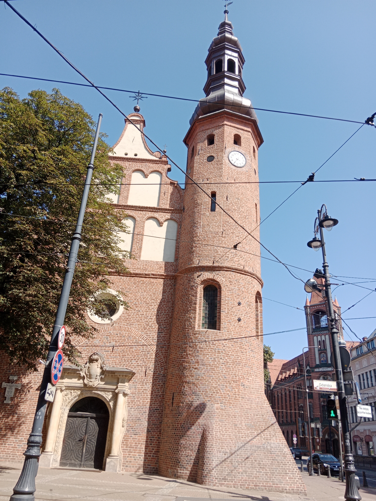 The image captures a low-angle view of the brick facade of a church in Bydgoszcz, Poland. The church features a tall, cylindrical tower with a copper-clad spire. A clock face is visible on the tower, and several arched windows punctuate the brickwork. To the left of the tower, a section of the church with stepped gables and decorative arched niches is visible. An ornate, arched doorway with heavy wooden doors is situated at the base of this section. A mature tree with dense green foliage stands to the left of the church entrance. In the foreground, a dark gray lamppost with multiple light fixtures is prominent, along with overhead electrical wires crisscrossing the sky. Traffic signs are attached to the lamppost, including a circular sign with a red border and a '30' inside, and a blue rectangular sign with a red border and a white cross. The sky is clear and bright blue. In the background, another church tower with a red roof and arched openings is visible, along with glimpses of urban buildings and a street with parked cars. A traffic light showing a green signal is also present. The overall scene suggests a sunny day in an urban setting.