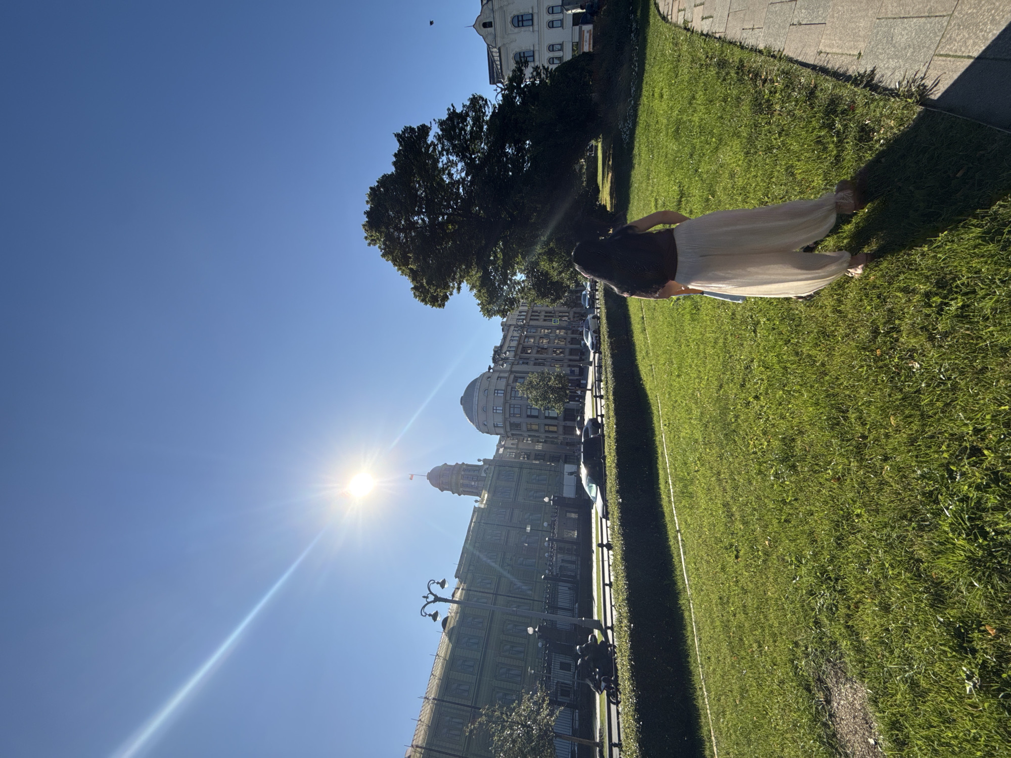 A woman stands in a grassy park area, her back to the camera, looking towards a large, dark green tree on her right. The tree is in full leaf and casts a long shadow across the grass. Behind the tree, a light-colored building with white columns is visible.
In the background, a street runs horizontally across the frame, with several cars and a motorcycle visible. On the other side of the street is a long, pale green building with scaffolding on its left side. The building has many windows and a domed tower with a flag on top. Further down the street, another domed building is visible. The sun is shining brightly, creating a lens flare in the upper left quadrant of the image and casting long shadows across the grass. The sky is a clear, bright blue. The location appears to be a public space within a city, possibly a park or square adjacent to historical buildings.