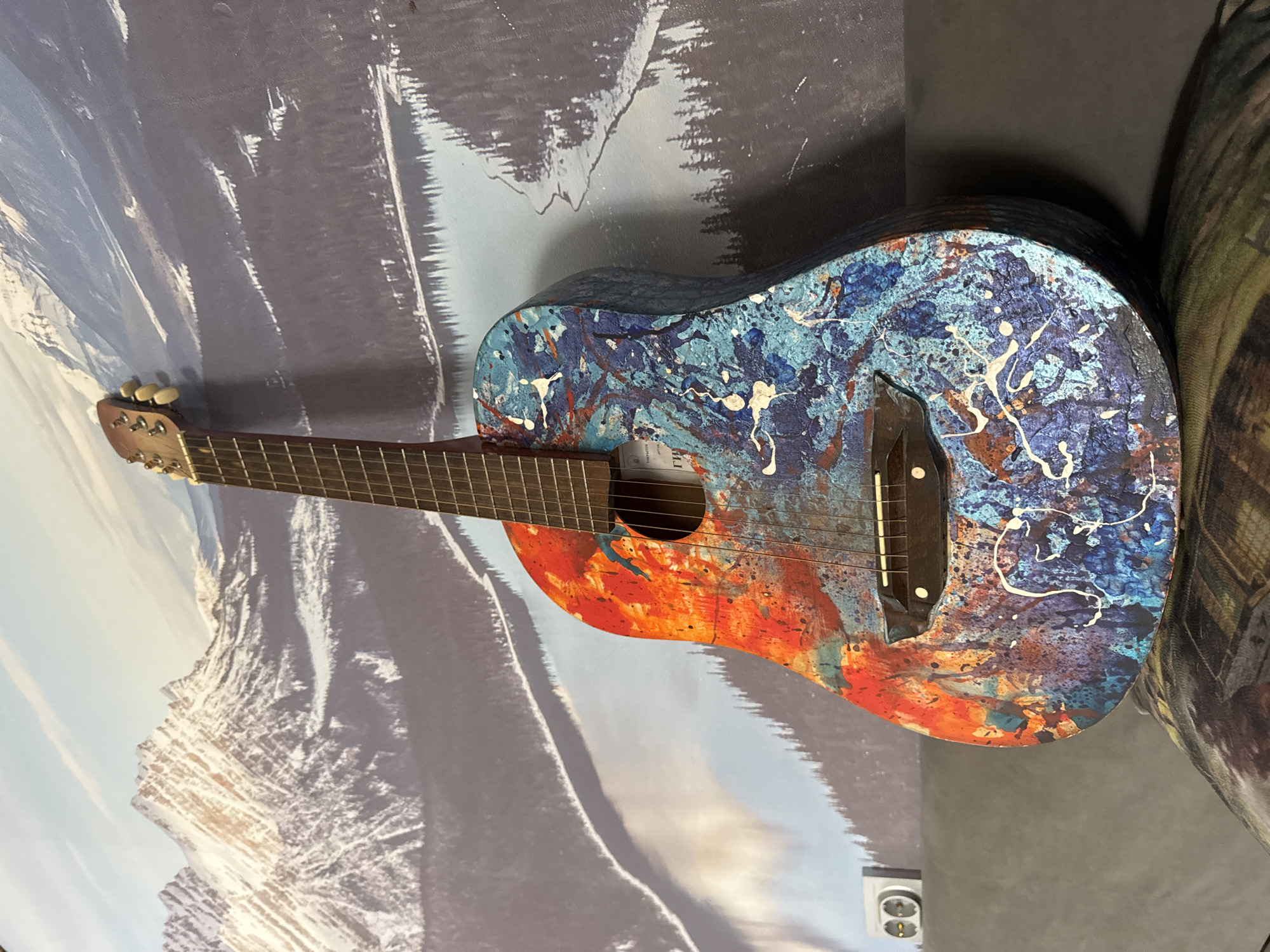A horizontally oriented, close-up shot shows a decorated acoustic guitar resting against a wall. The body of the guitar is brightly painted in shades of blue and orange, with white splatters. The guitar has a dark brown neck and headstock with light-colored tuning pegs.
The guitar is positioned in front of a wall mural depicting a mountain range under a sky with clouds. To the right of the guitar, there is a textured, grey or green surface, possibly a portion of a piece of furniture.
In the lower right corner of the image, an electrical outlet can be seen, indicating the scene is likely indoors. The location is in the city of Chaikovsky, Russia.