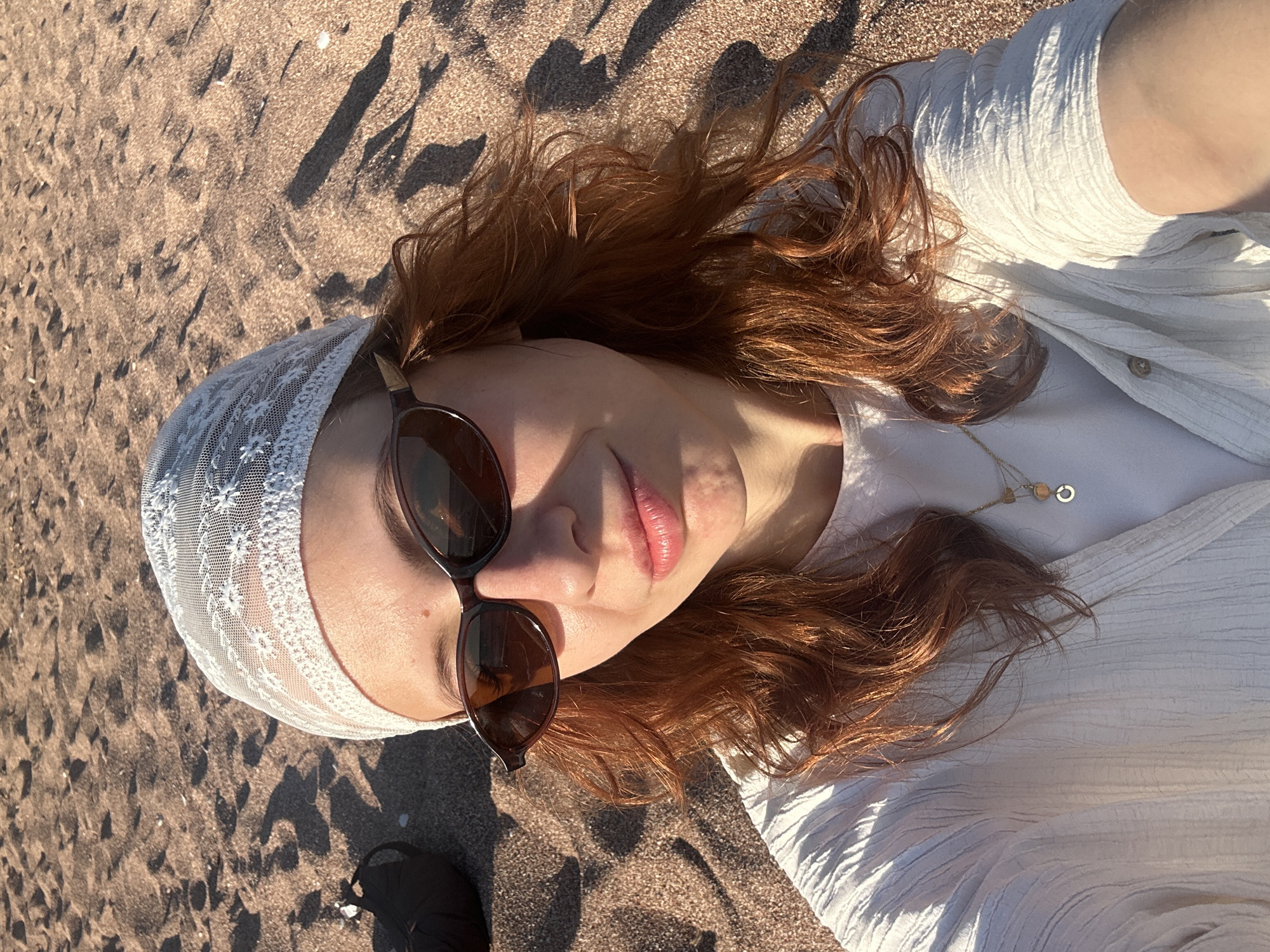 The image shows a woman lying on a beach. She has red hair and is wearing a white lace headscarf and sunglasses. She is wearing a white shirt and a necklace. The sand is brown. The location is Petrovac centar, Montenegro.