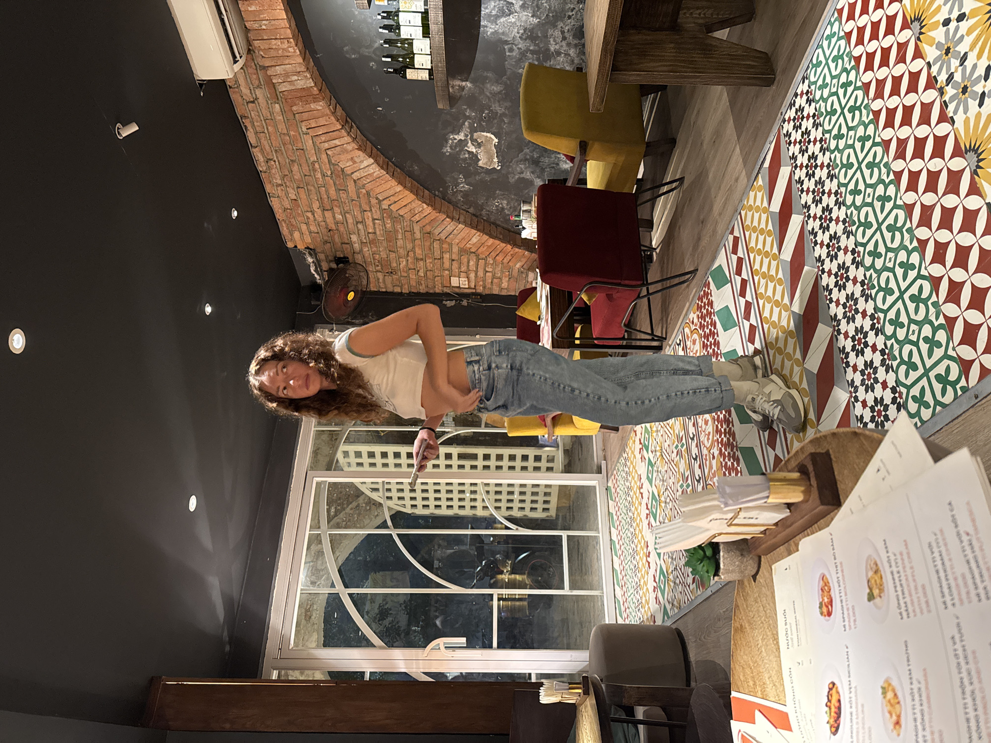 This image shows a young woman with curly brown hair standing in a restaurant. She is wearing a white crop top and blue jeans. The restaurant has a unique interior design with exposed brick walls, colorful patterned tiles on the floor, and modern furniture. There are yellow and red chairs visible, as well as a wooden table. A large window with intricate white panes is also a prominent feature. The woman appears to be holding a fork in her right hand and looking towards the camera with a smile. Menus are laid out on a wooden table in the foreground. The lighting suggests it could be evening or indoors with artificial lighting. The visible text on the menus includes Vietnamese food items like "ME SPAGHETTI KOT THIT BO BAM" and "NƯỚC SUỐI". The overall atmosphere seems casual and inviting, possibly a cozy dining establishment in Hanoi, Vietnam.